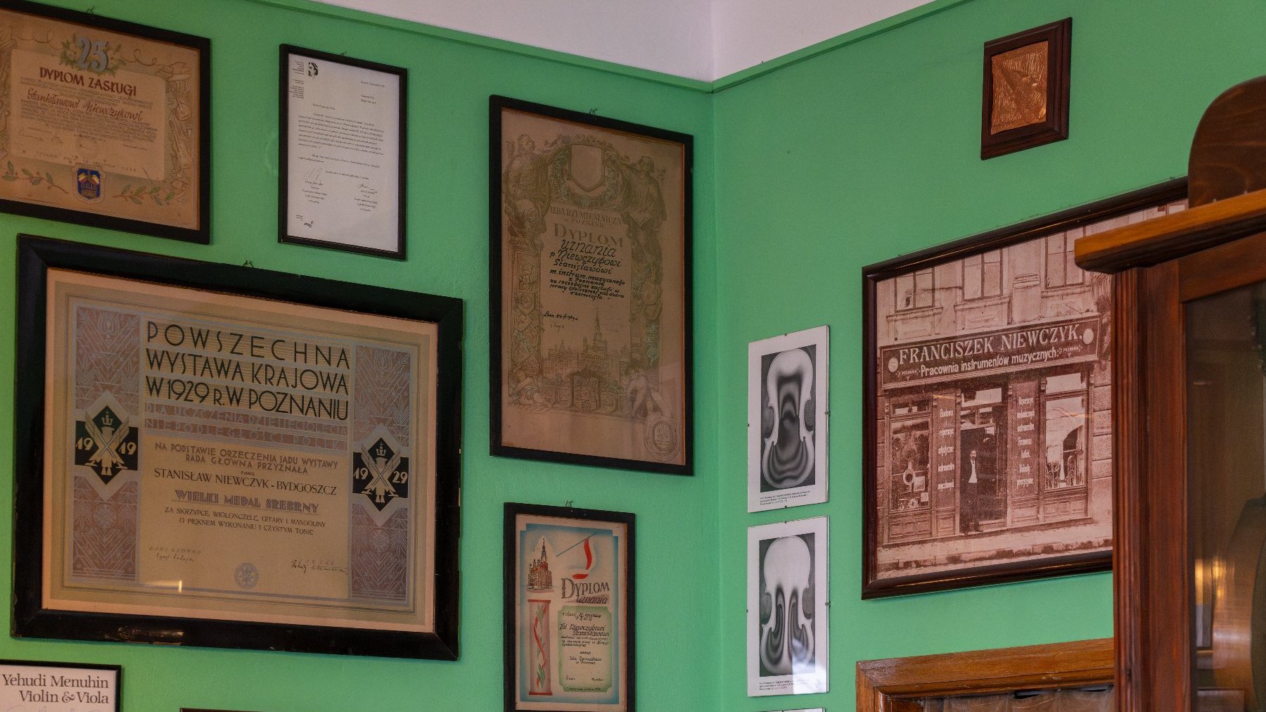 Zielona ściana w warsztacie lutniczym, na której wiszą liczne dyplomy i certyfikaty w ramach, w tym duży dyplom z Powszechnej Wystawy Krajowej w 1929 roku w Poznaniu. Na ścianie wisi także oprawiona w ramę historyczna wizytówka z napisem: "FRANCISZEK NIEWCZYK Pracownia instrumentów muzycznych".