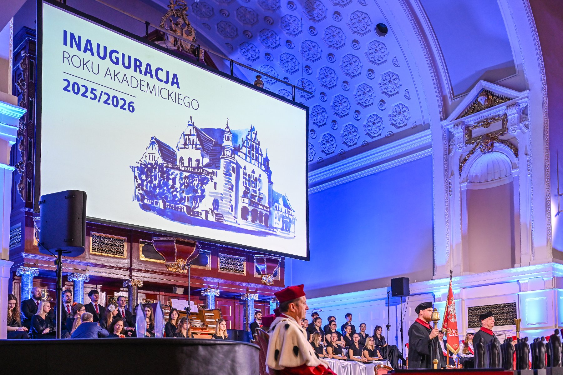 Zdjęcie przedstawia ekran z napisem "inauguracja roku akademickiego" i stojących pod nim ludzi. - grafika artykułu
