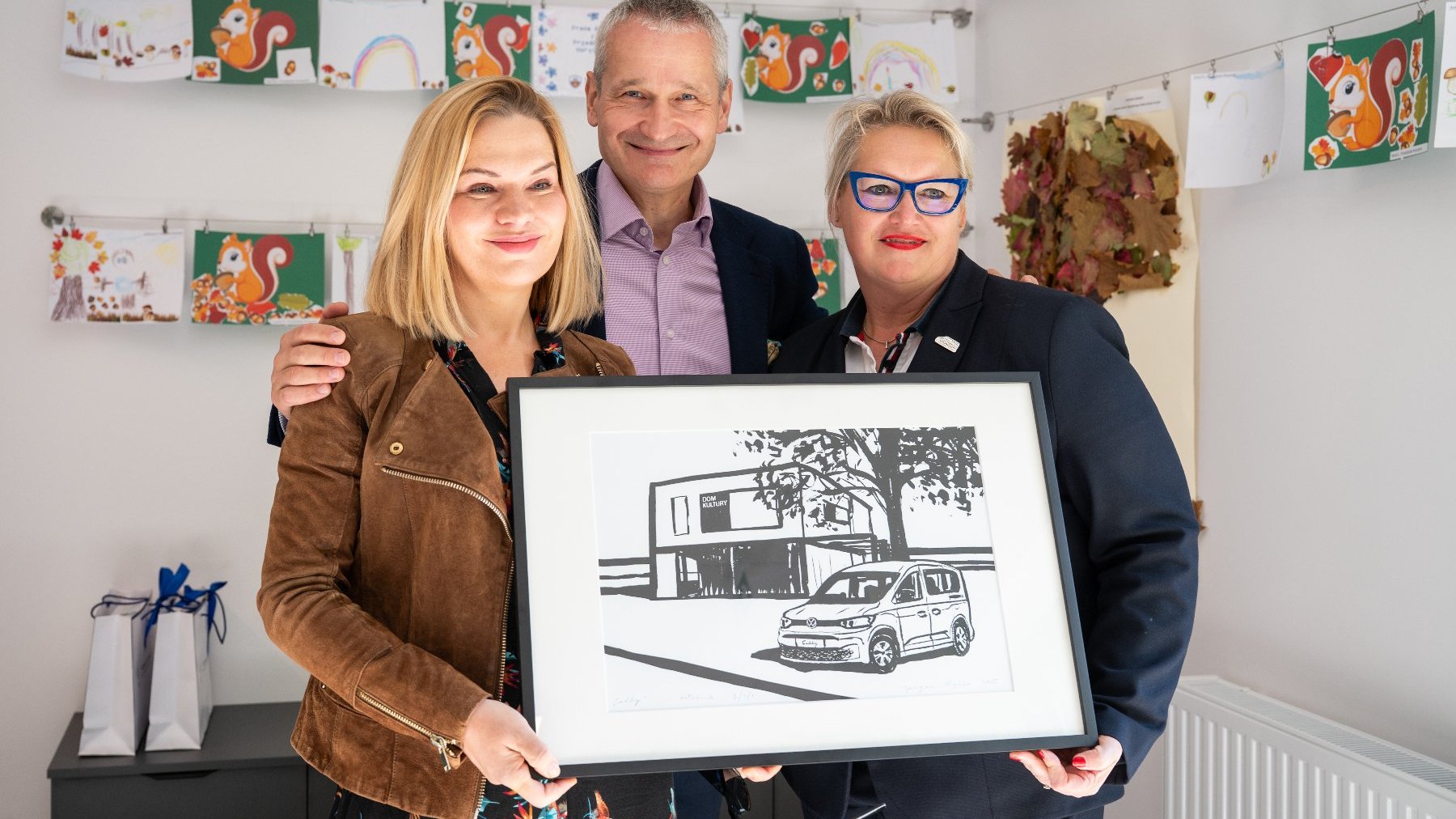 Trzy osoby (dwie kobiety i mężczyzna) stoją w jasnym pomieszczeniu z dziecięcymi rysunkami wiewiórek na ścianie. Wszyscy trzymają oprawioną w ramę czarno-białą grafikę przedstawiającą nowoczesny, dwupiętrowy budynek oraz samochód typu van. Kobieta po prawej ma niebieskie okulary i krótko ścięte włosy, a kobieta po lewej jest ubrana w brązową kurtkę zamszową.