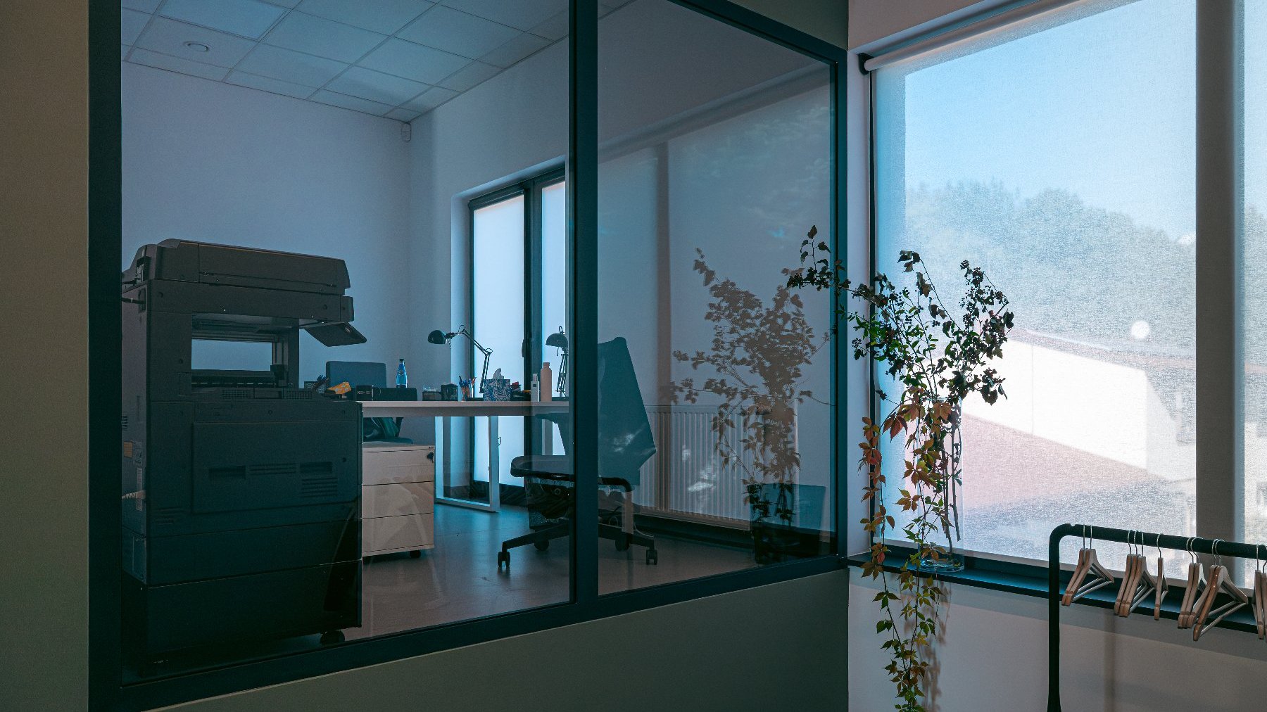 Wnętrze nowoczesnego biura z przeszkloną, czarną ścianą działową. Przez szkło widoczne jest drugie pomieszczenie biurowe z drukarką/kopiarką, biurkiem, krzesłem i oknem w tle. Po prawej stronie widać duże okno z opuszczoną, białą roletą, a na parapecie stoi wazon z roślinami. Na pierwszym planie widoczny jest wieszak na ubrania.