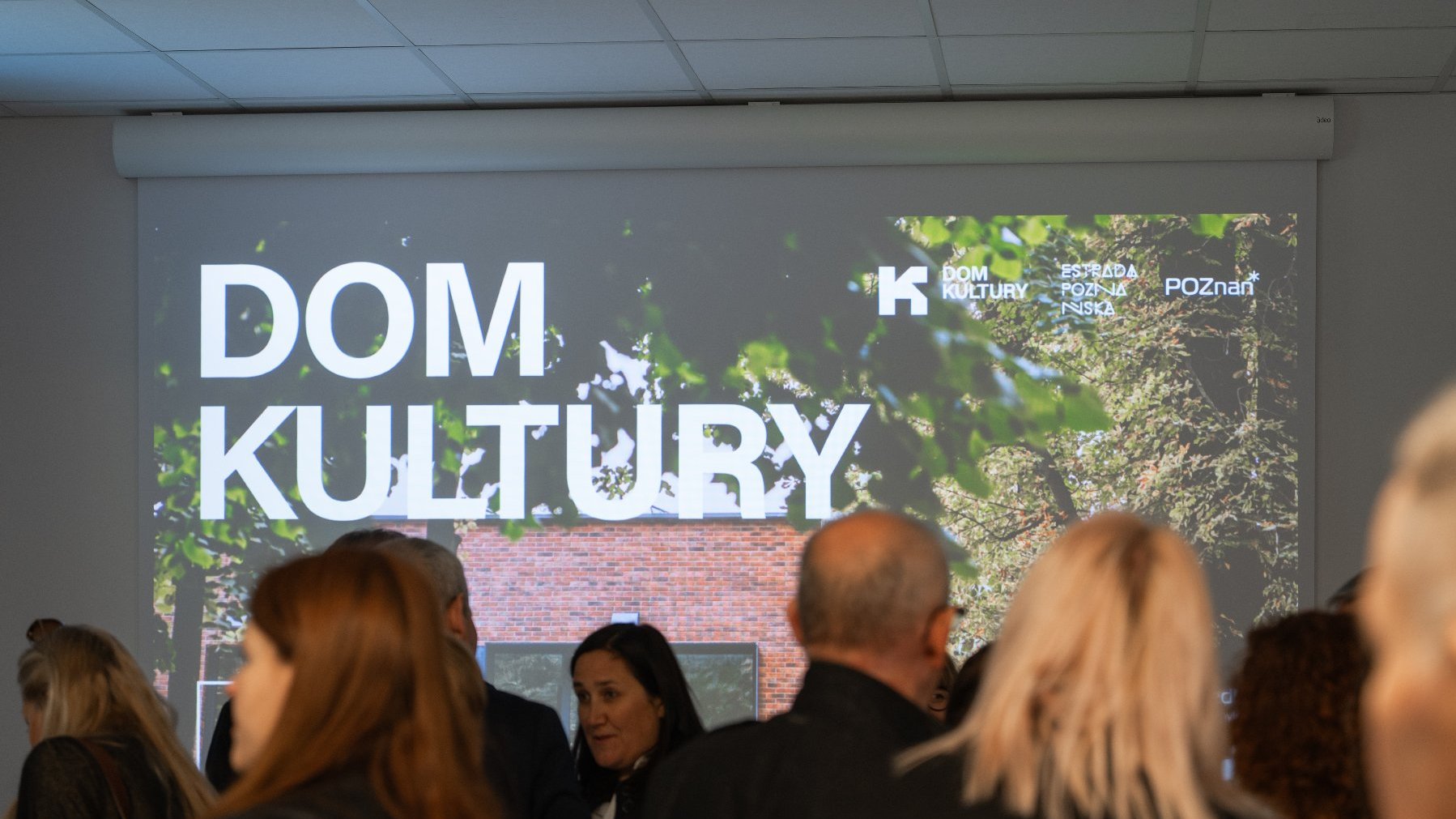 Ekran projekcyjny w jasnym pomieszczeniu z wyświetloną grafiką z napisem: "DOM KULTURY" dużą, białą czcionką, na tle liści i cegły. W tle, u góry ekranu, widoczne są mniejsze loga: "K Estrada Poznańska" i "POZnań". Na pierwszym planie widoczny jest tłum publiczności (widziany od tyłu) zgromadzony na wydarzeniu.