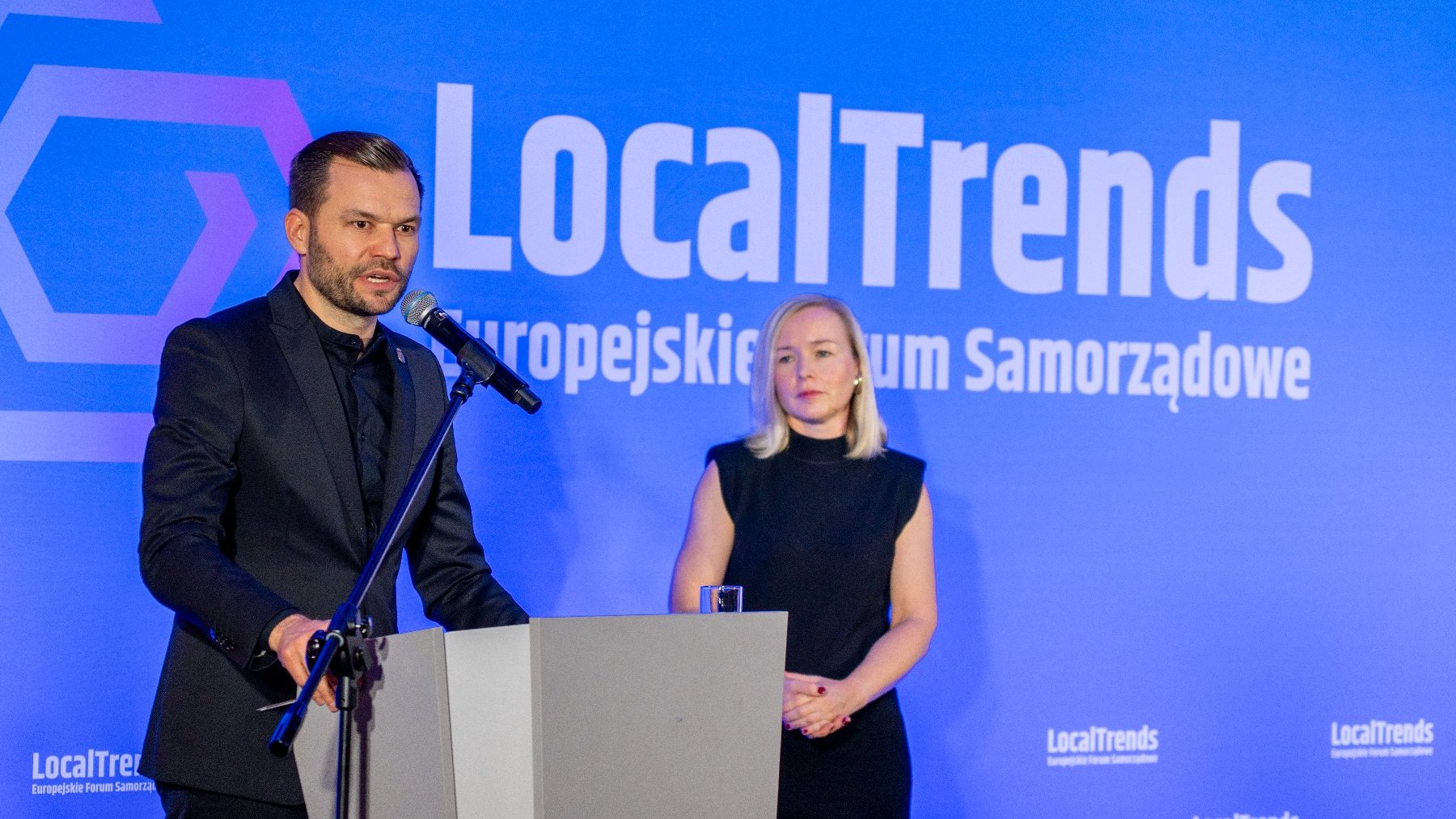 Mężczyzna w czarnym garniturze przemawia z szarej mównicy, trzymając ręce na pulpicie. Obok niego stoi kobieta w czarnej sukience bez rękawów. W tle niebieski ekran z napisem: "LocalTrends Europejskie Forum Samorządowe".