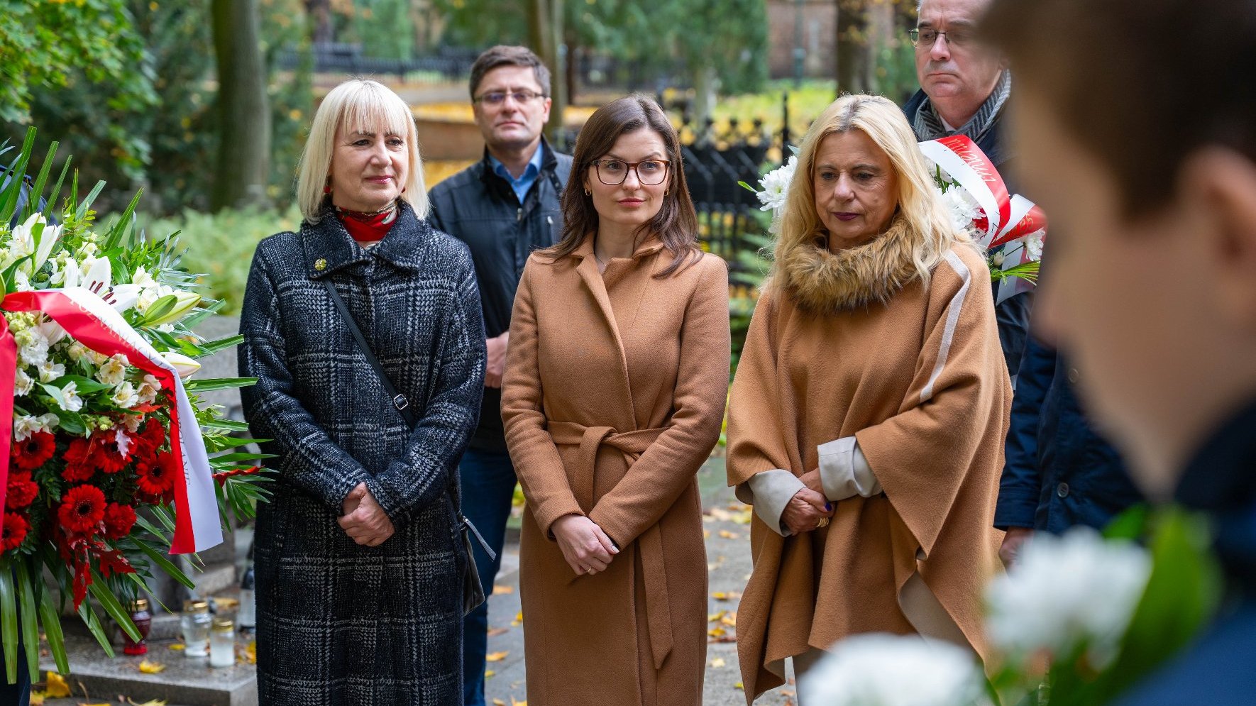 Grupa osób stoi na cmentarzu. Na pierwszym planie trzy kobiety: w czarnym płaszczu w kratę, w brązowym płaszczu i w beżowym ponczo z futrzanym kołnierzem. Za nimi stoją dwaj mężczyźni. Po lewej stronie widoczny duży bukiet z biało-czerwonych kwiatów.