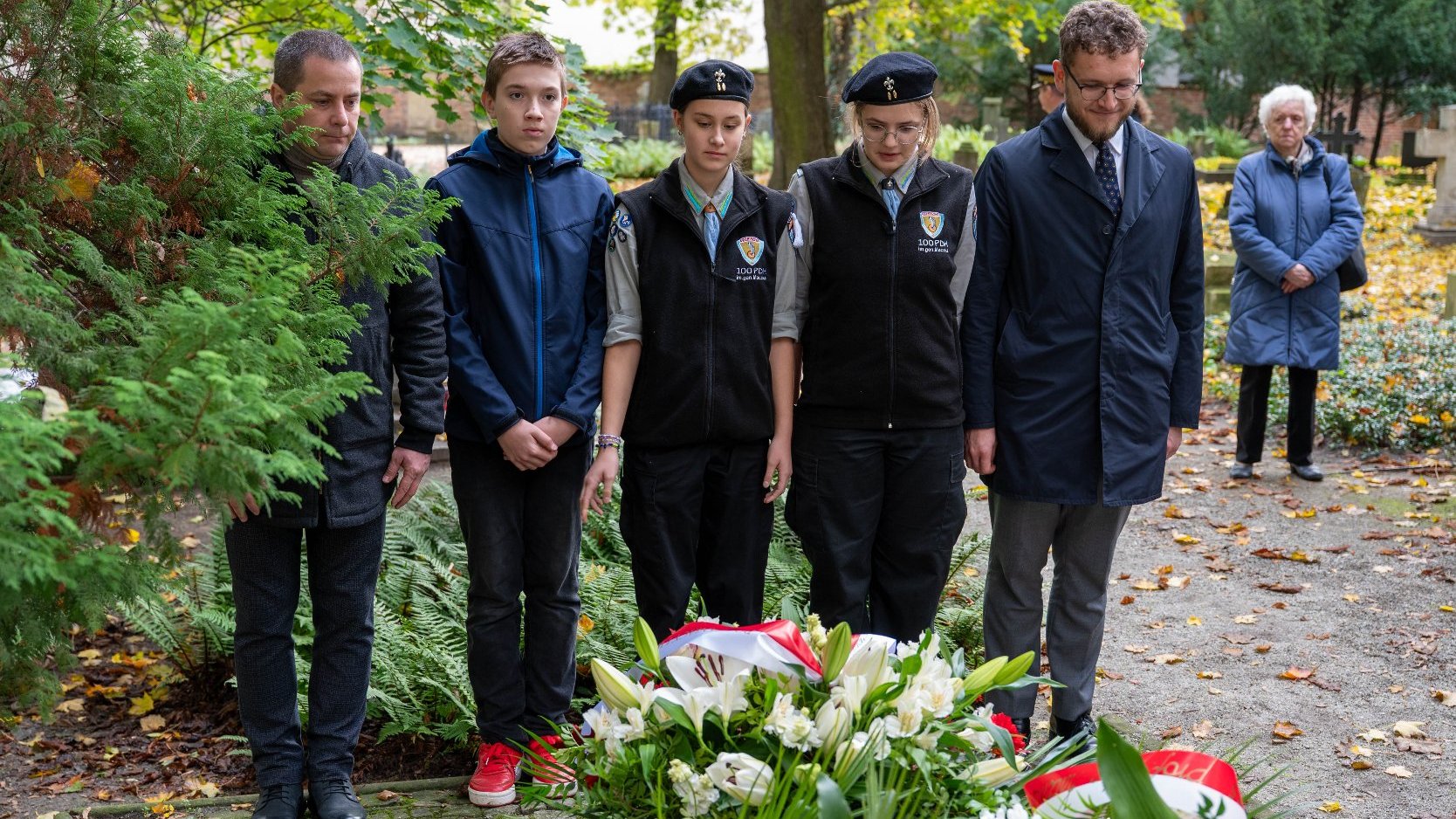 Grupa pięciu osób stoi w skupieniu nad grobem. Wśród nich dwoje młodych ludzi w czarnych mundurach harcerskich. Przed nimi duży wieniec z białych lilii i czerwonych goździków z biało-czerwoną szarfą. W tle stoi starsza kobieta i jesienna zieleń.