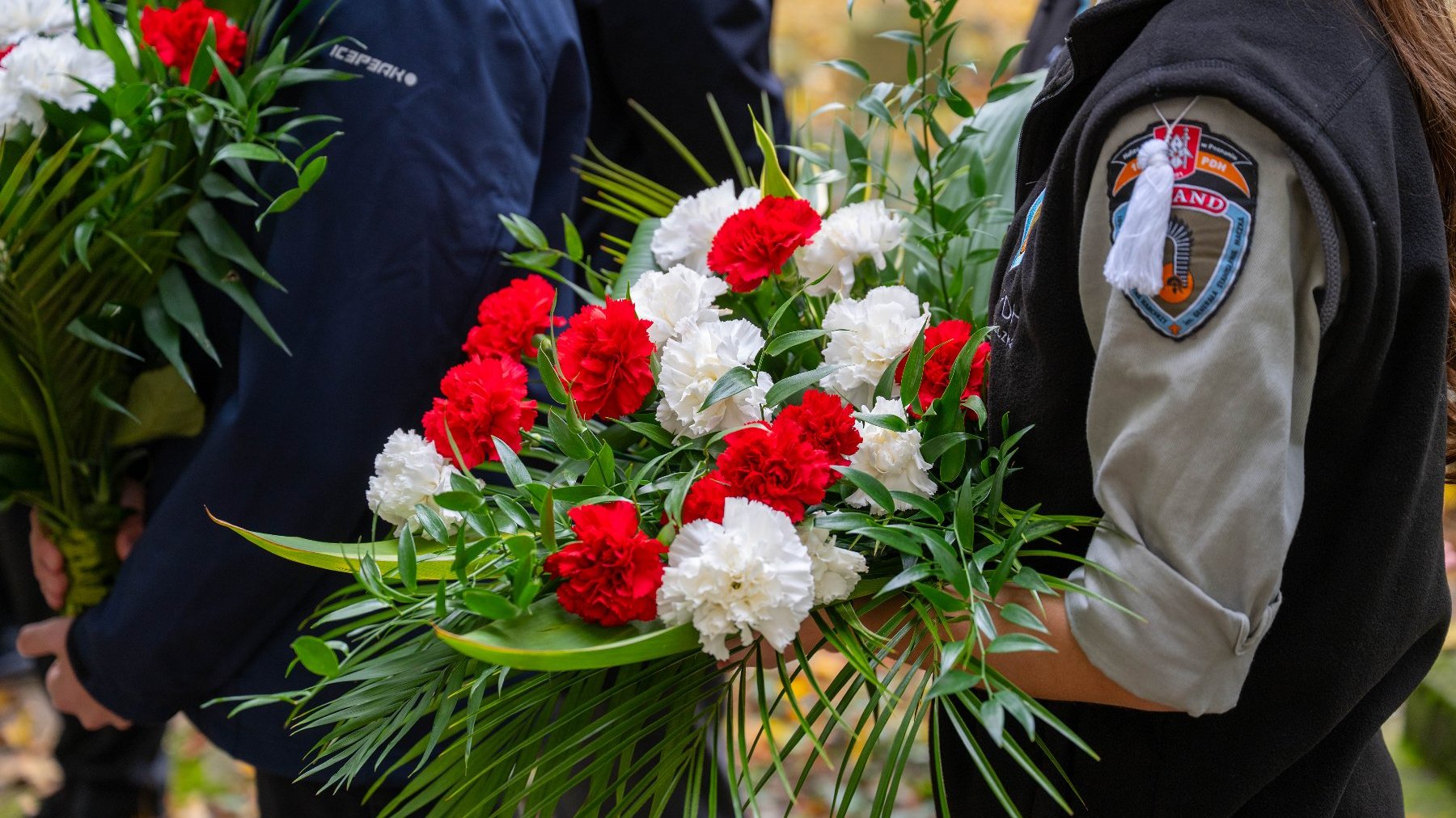 Zbliżenie na bukiety trzymane przez dwie osoby. Bukiety składają się z czerwonych i białych goździków oraz zielonych liści. Po prawej widoczny fragment rękawa w czarnej kamizelce z naszywką.