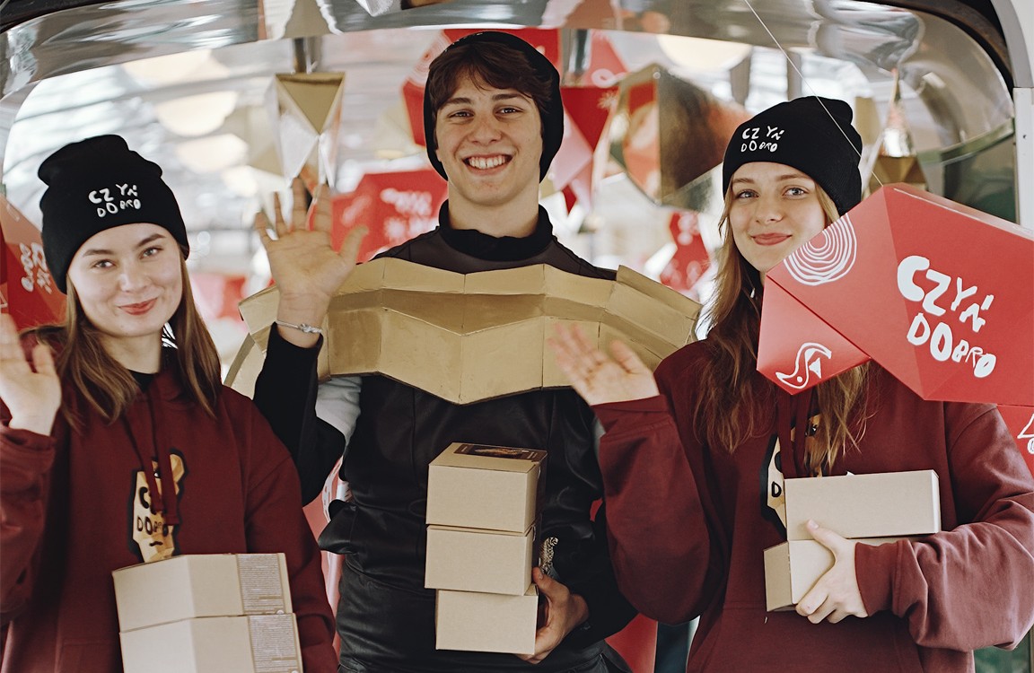 Na zdjeciu grupa uśmiechniętych ludzi, trzymających pudełka z rogalami, w tramwaju ozdobionym papierowymi rogalami - grafika artykułu