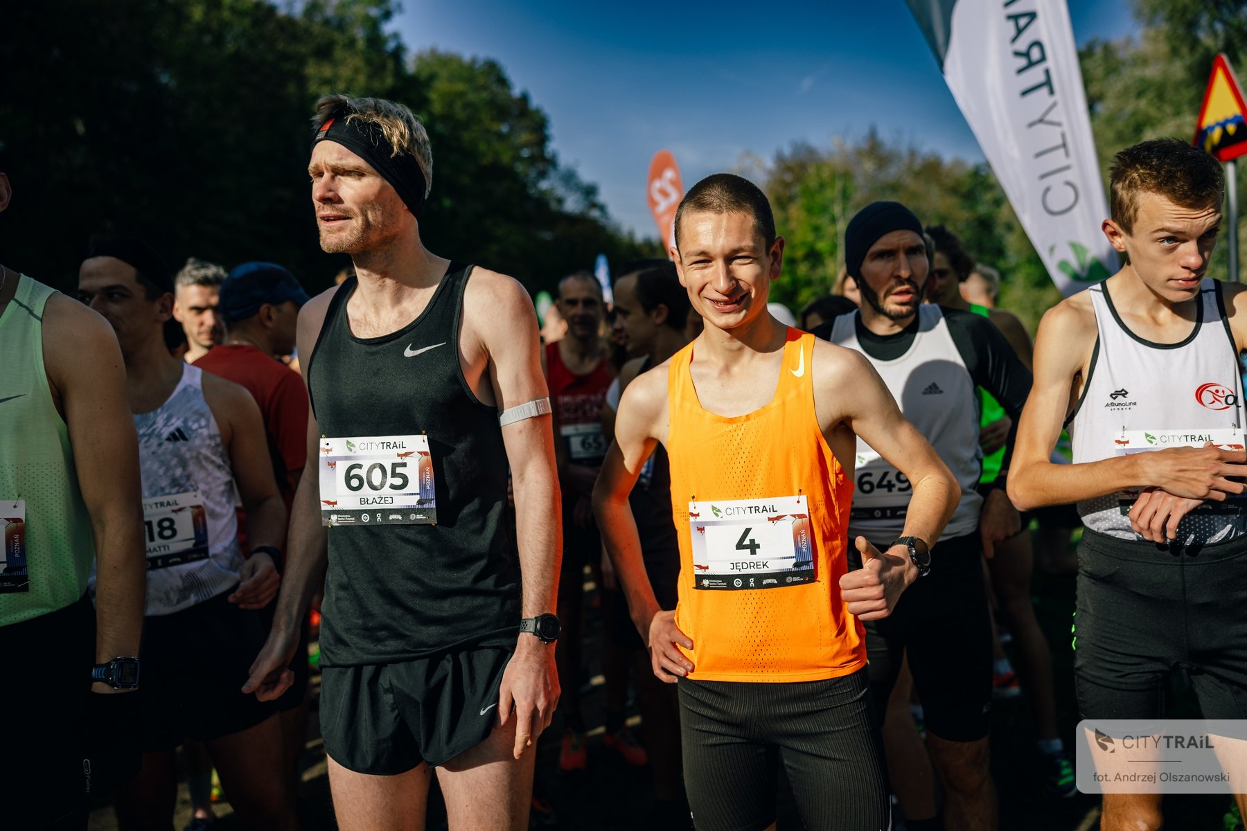 Grupa biegaczy stoi na starcie zawodów, czekając na rozpoczęcie biegu. Na pierwszym planie dwóch mężczyzn w sportowych koszulkach: jeden w czarnej z numerem 605, drugi w pomarańczowej z numerem 4, uśmiecha się. - grafika artykułu