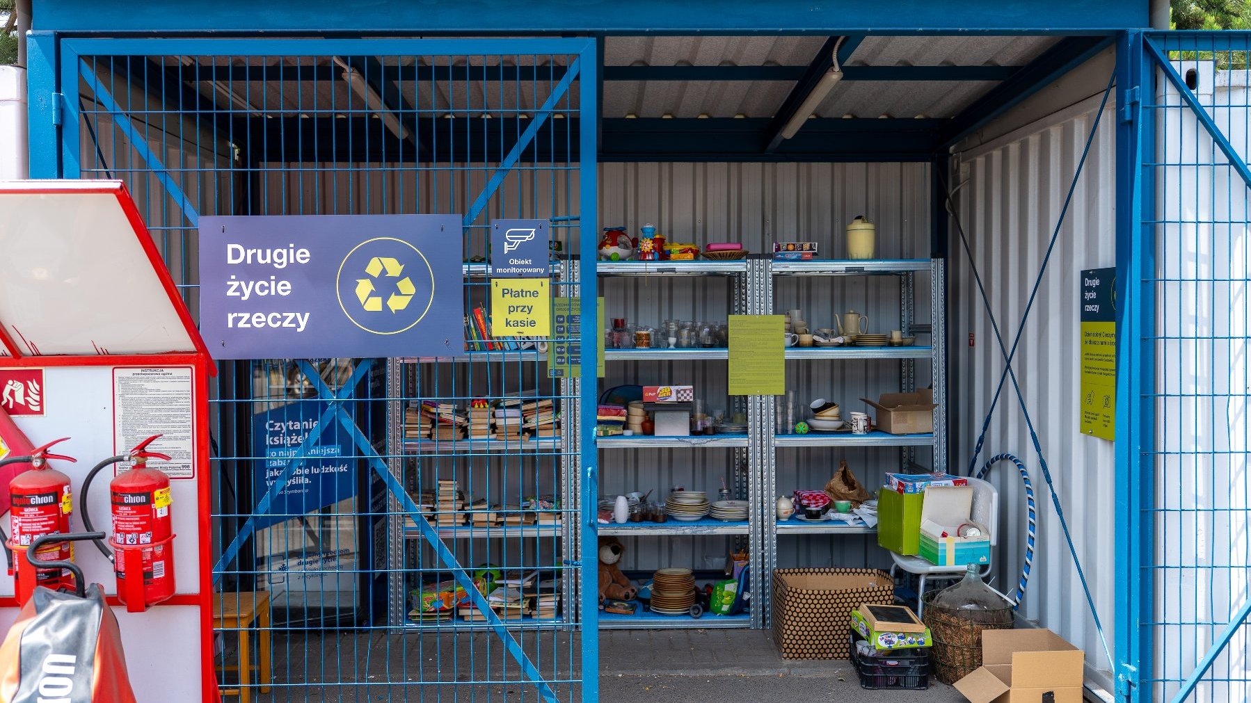 Zdjęcie przedstawia półkę, na której znajdują się książki, zabawki, ozdoby. Półka znajduje się w pomieszczeniu za bramą. brama jest otwarta.