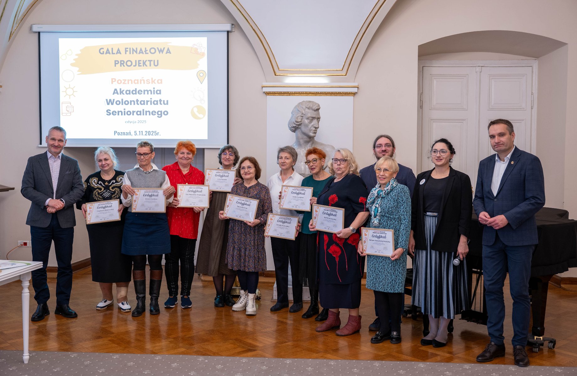 Grupa osób pozuje do zdjęcia w sali z drewnianą podłogą, podczas Gali Finałowej. W centrum ujęcia w rzędzie stoi dziesięć starszych kobiet, trzymających certyfikaty, z uśmiechem skierowanym w stronę aparatu. - grafika artykułu
