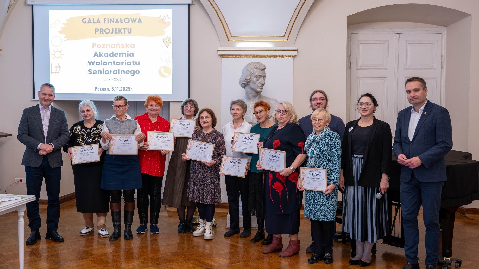 Grupa osób pozuje do zdjęcia w sali z drewnianą podłogą, podczas Gali Finałowej. W centrum ujęcia w rzędzie stoi dziesięć starszych kobiet, trzymających certyfikaty, z uśmiechem skierowanym w stronę aparatu.