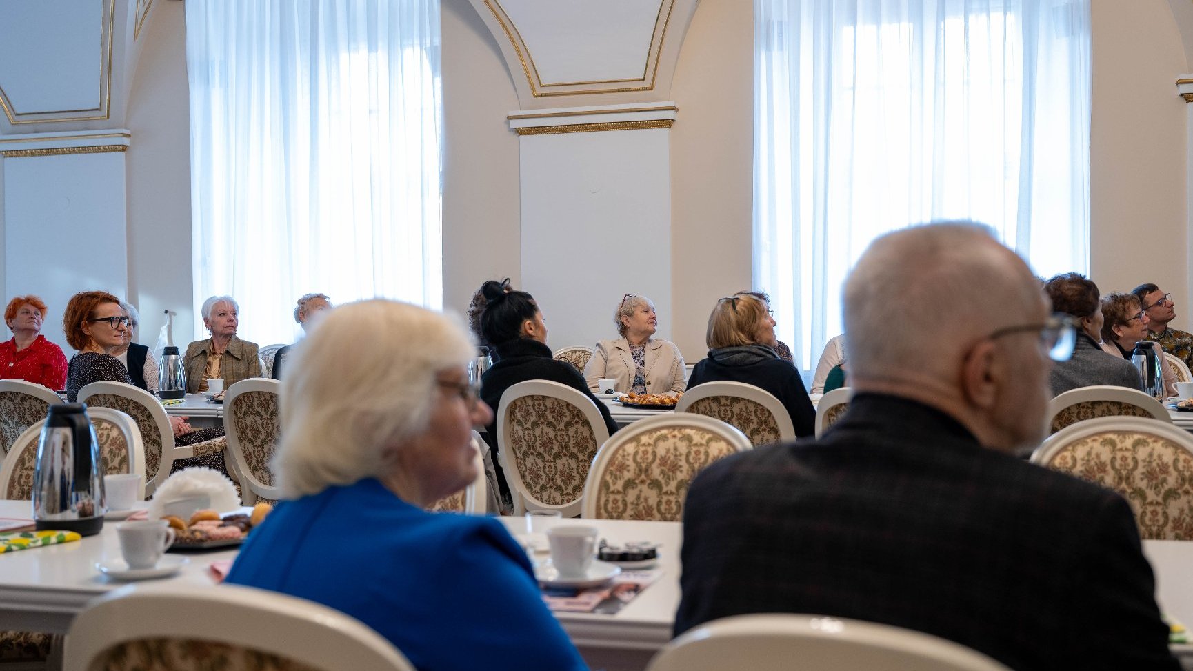 Uczestnicy spotkania lub konferencji siedzą przy długim stole, odwróceni plecami do aparatu lub siedzący z boku. Na stole stoją filiżanki z kawą i talerzyki. W tle widoczne wysokie okna zasłonięte białymi, długimi firanami.