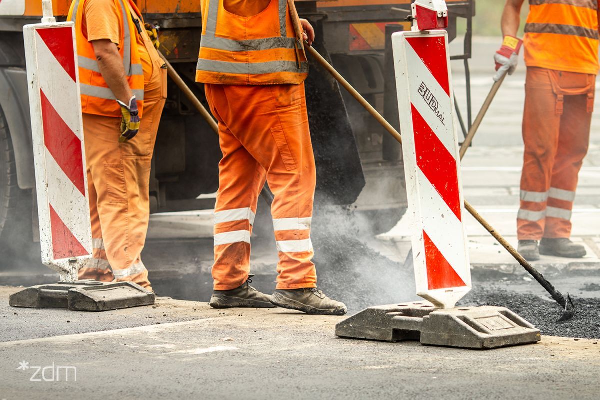 Fragment budowy drogi. Trzech pracowników ubranych w pomarańczowe spodnie i kamizelki odblaskowe stoi na asfalcie, trzymając szpadle lub grabie do wyrównywania nawierzchni. Na pierwszym planie dwa biało-czerwone słupki drogowe. Z lewej strony na dole widoczne małe, białe logo w cudzysłowie: "*zdm". - grafika artykułu