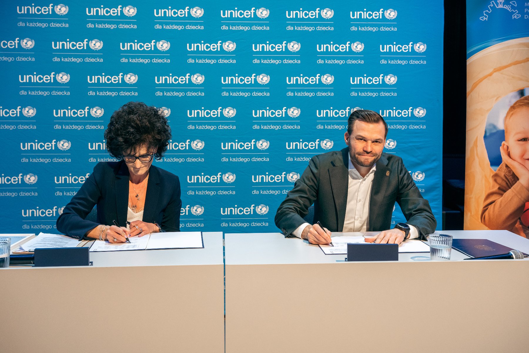Na zdjęciu kobieta i mężczyzna podpisujący dokumenty, za nimi ścianka z logo UNICEF - grafika artykułu
