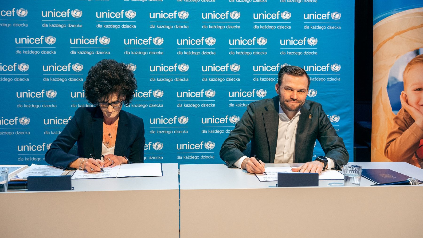 Na zdjęciu kobieta i mężczyzna podpisujący dokumenty, za nimi ścianka z logo UNICEF