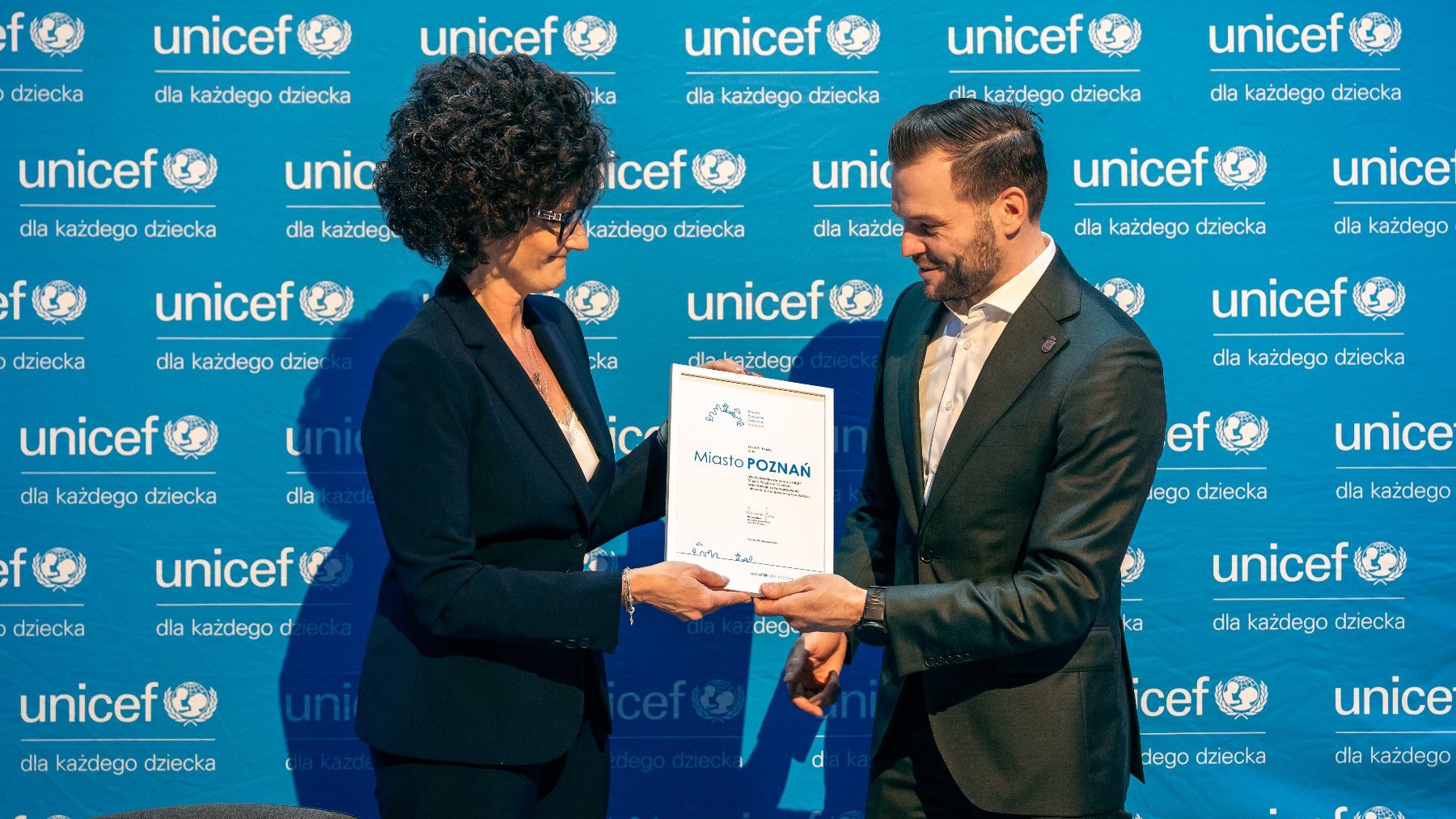 Na zdjęciu kobieta i mężczyzna, przekazują sobie certyfikat na tle ścianki z logo Unicef