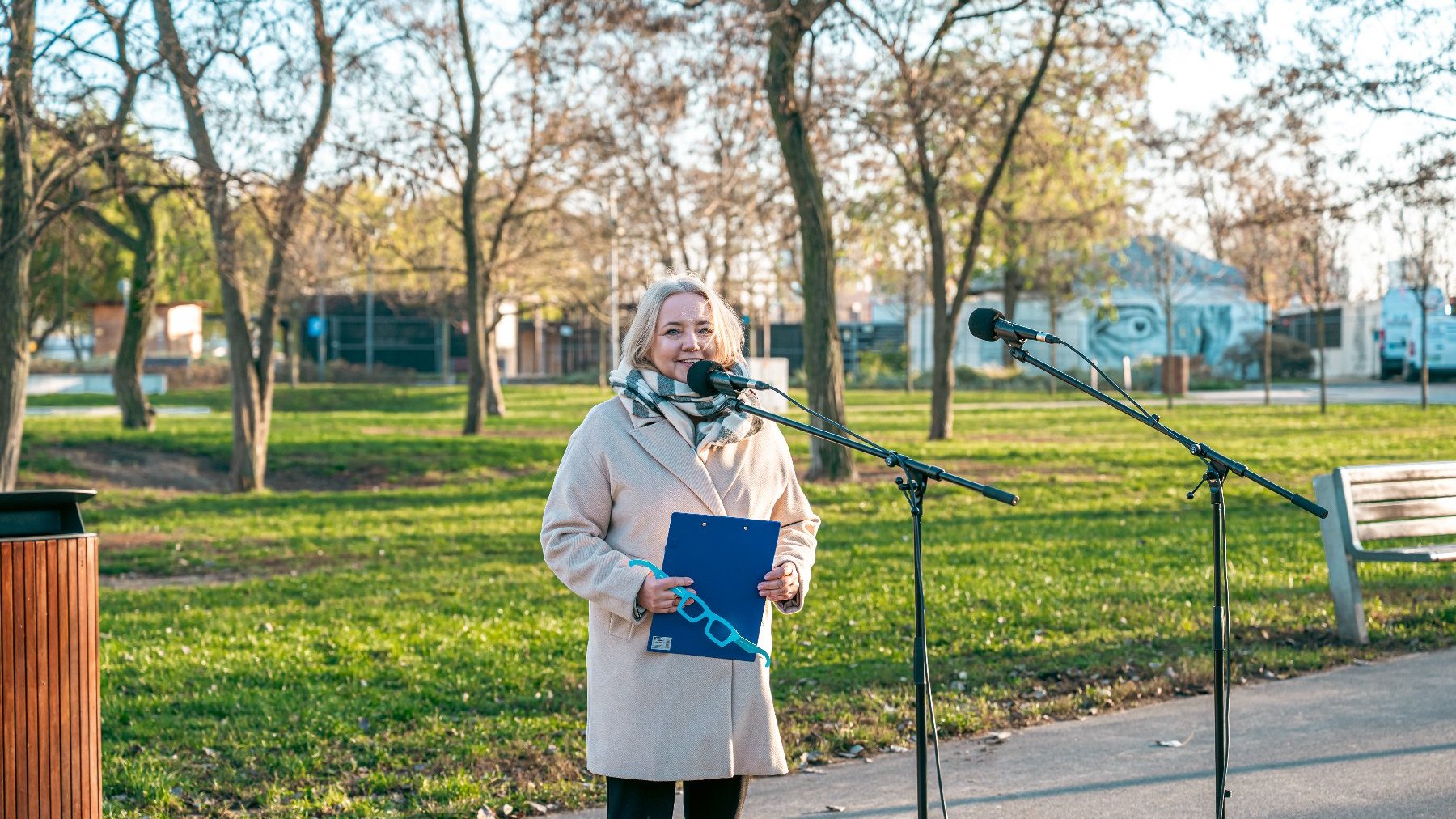Na zdjęciu kobieta mówiąca coś do mikrofonu, stojąca w przestrzeni parku