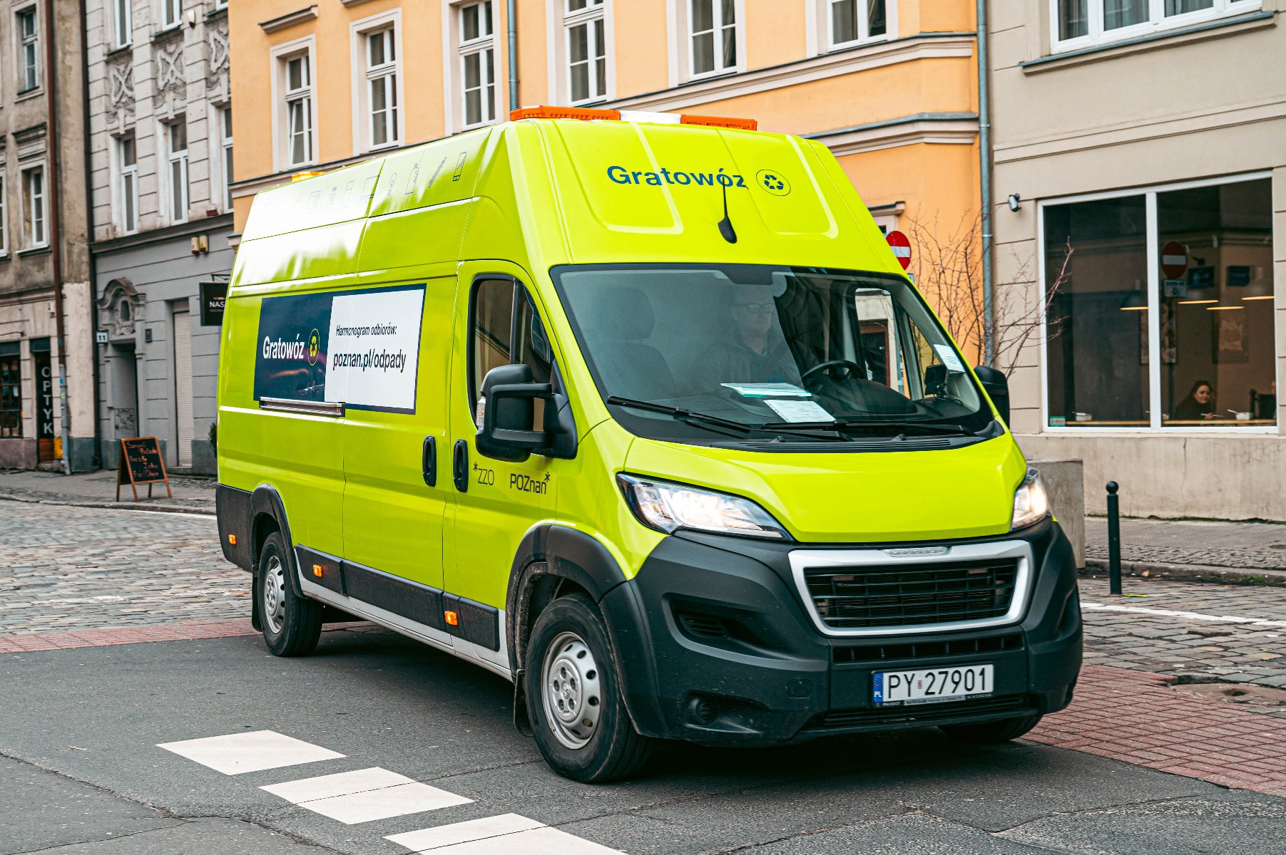Dostawczy samochód marki Peugeot w jaskrawożółtym, seledynowym kolorze, jadący po brukowanej ulicy w mieście. Na dachu samochodu pomarańczowe lampy i duży, zielony napis "Gratowóz". Po bokach reklama i adres strony, w tym: "poznan.pl/odpady". - grafika artykułu