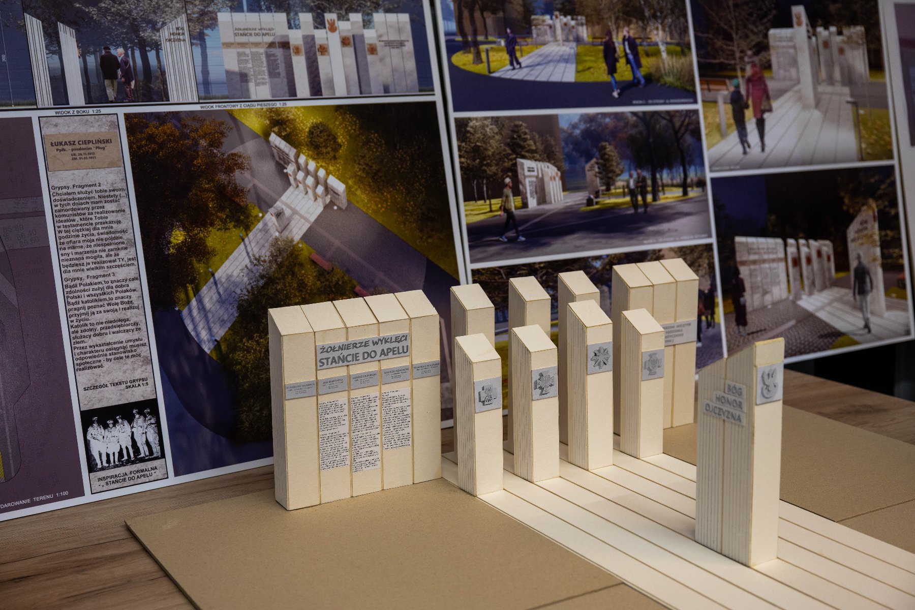 Na drewnianym stole leży plansza z wizualizacjami i tekstem, a przed nią stoi przestrzenna makieta pomnika. Makieta składa się z kilku beżowych prostopadłościanów symbolizujących kamienne tablice. - grafika artykułu