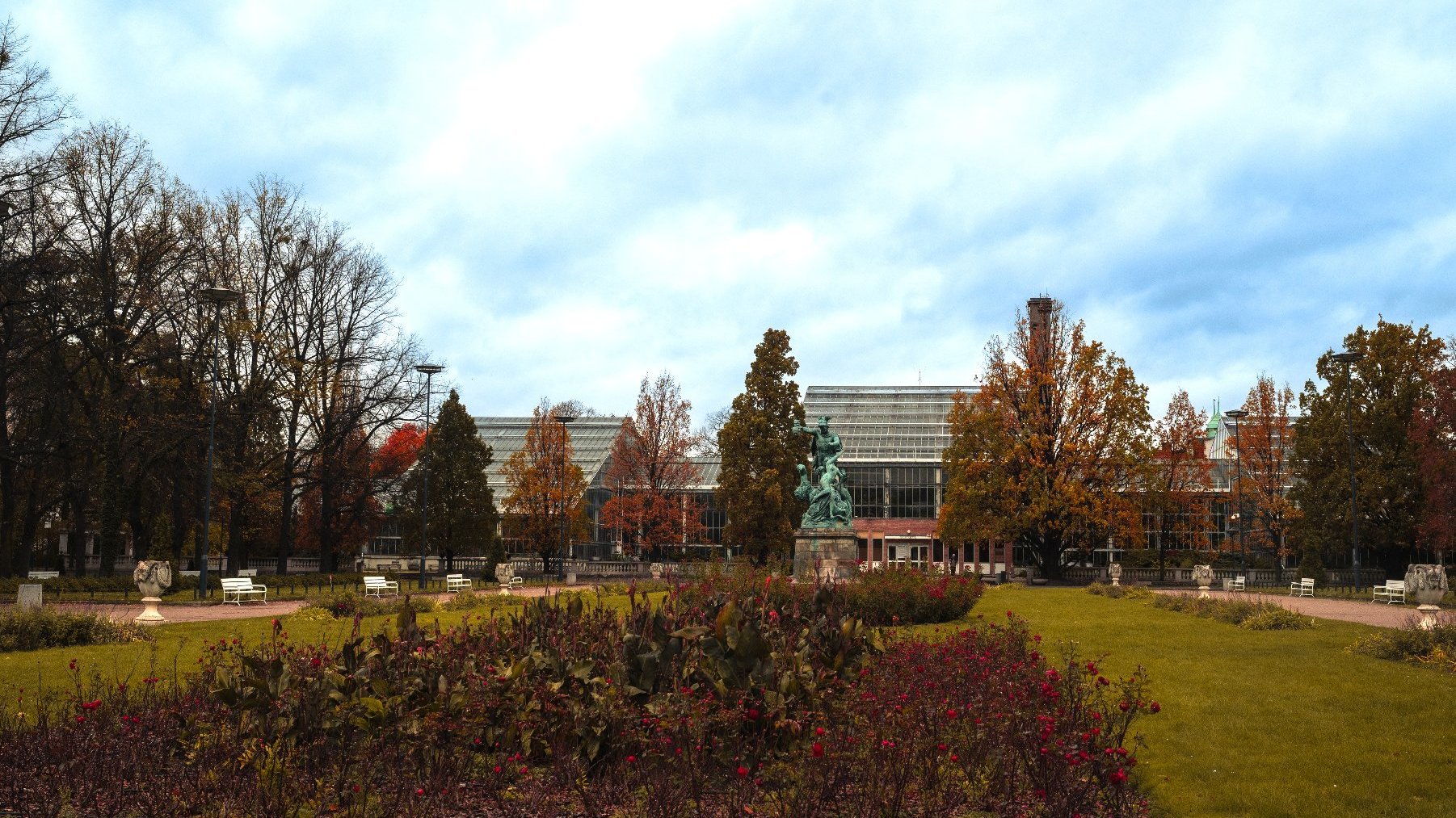 Zdjęcie przedstawia budynek palmiarni i otaczającą ją zieleń oraz stojacy obok pomnik.