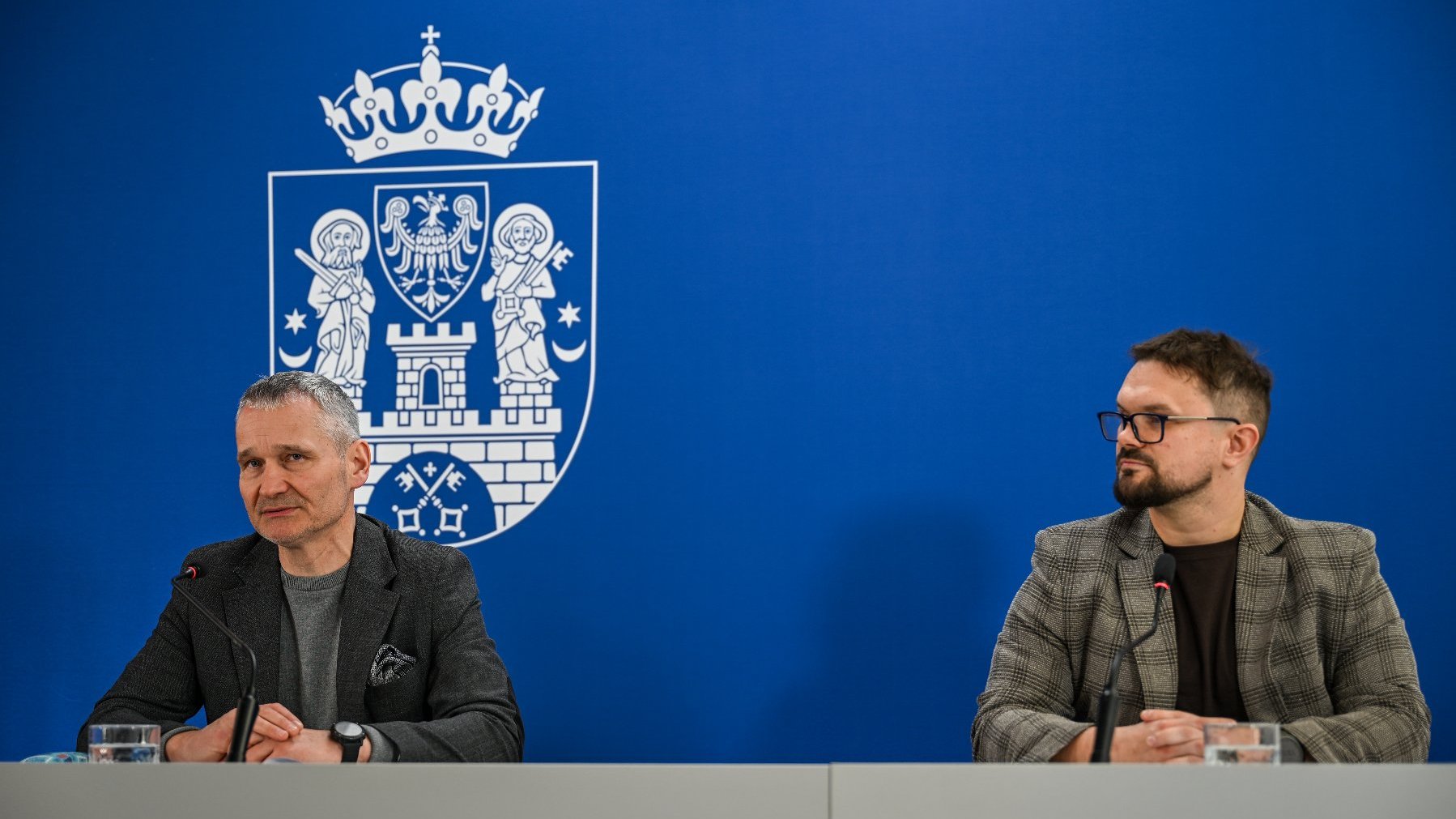 Na zdjęciu dwaj mężczyźni za stołem konferencyjnym, za nimi ścianka z herbem miasta