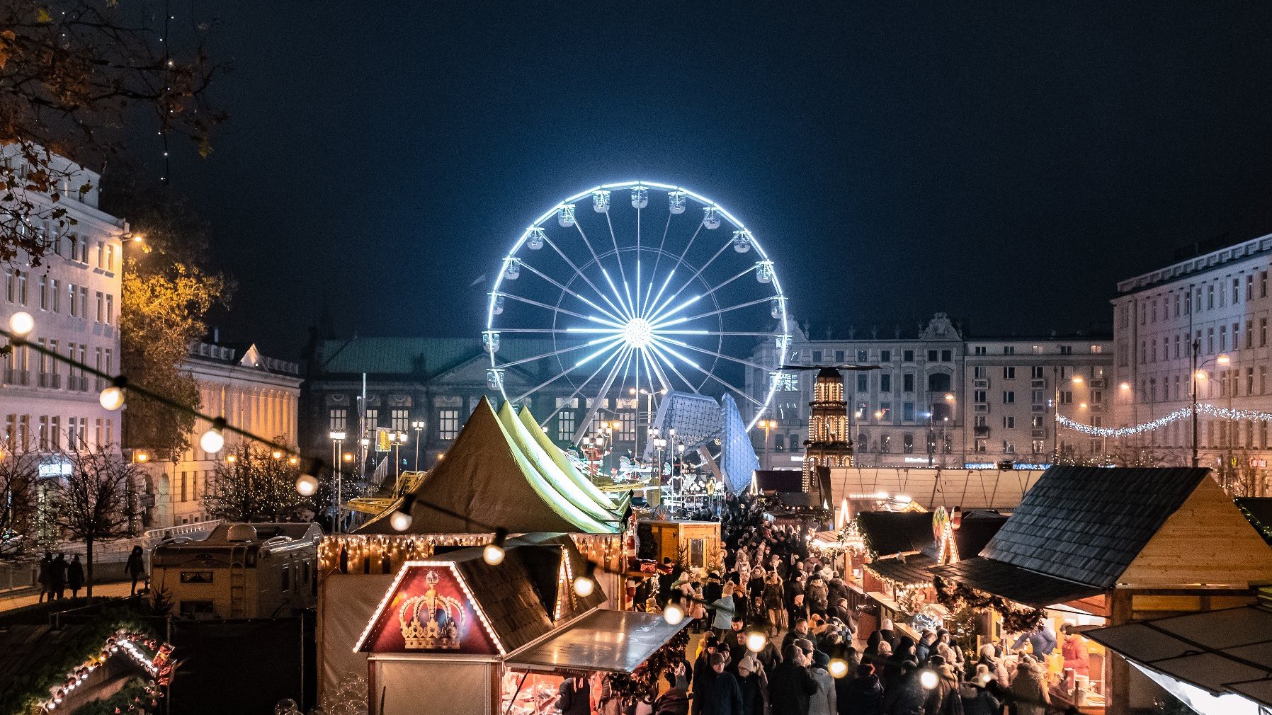 WIdok na Betlejem Poznańskie. Na pierwszym planie drewniane, rozświetlone budki. Między nimi spacerują ludzie ubrani w ciepłe kurtki, czapki i szaliki. W tle rozświetlony na biało Diabelski Młyn oraz stare kamienice. Niebo jest ciemne.