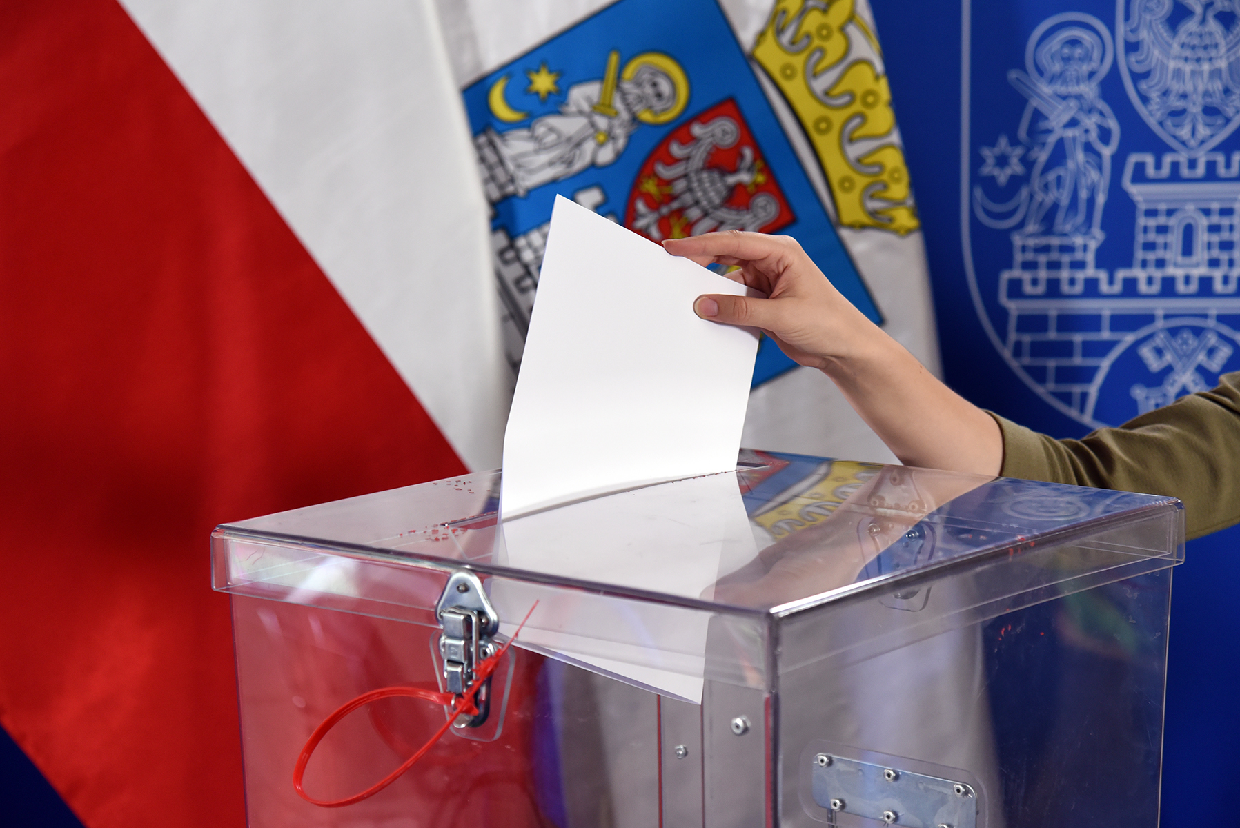 Na zdjęciu wyborca wrzuca kartę do głosowania do przezroczystej urny wyborczej. W tle widać flagi. - grafika artykułu