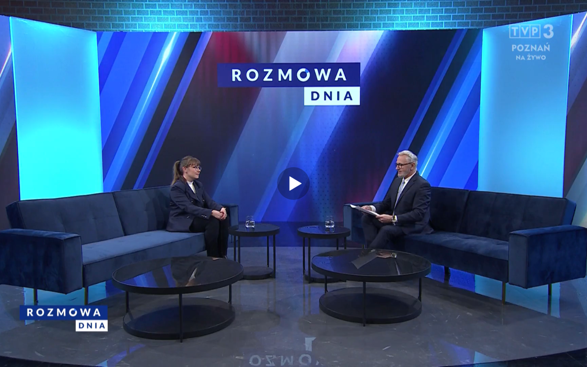 Dwie osoby siedzą na granatowych kanapach w studiu telewizyjnym programu "Rozmowa Dnia" na antenie TVP3 Poznań. Po lewej stronie kobieta w okularach, ubrana w ciemny żakiet i spodnie, z dłoniami złożonymi na kolanach. Po prawej stronie mężczyzna w garniturze, trzymający kartki. W tle nowoczesna scenografia w odcieniach niebieskiego z podświetlanymi panelami oraz logotyp programu. Na ekranie widoczne jest również oznaczenie "NA ŻYWO", wskazujące transmisję na żywo. - grafika artykułu