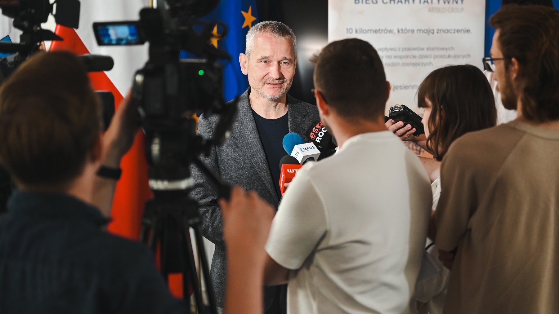 Na tle rollupu promującego Jesienny Bieg Charytatywny na rzecz Rodzicielstwa Zastępczego, stoi zastępca prezydenta Miasta Poznania. Dookoła stoję dziennikarze trzymający w rękach mikrofony.