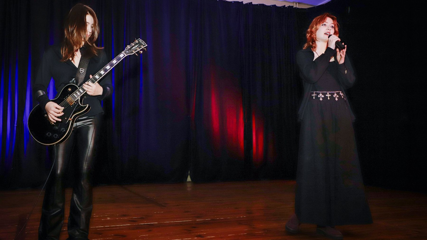 Występ artystyczny. Ciemne tło, na scenie kobieta z mikrofonem i mężczyzna z gitarą