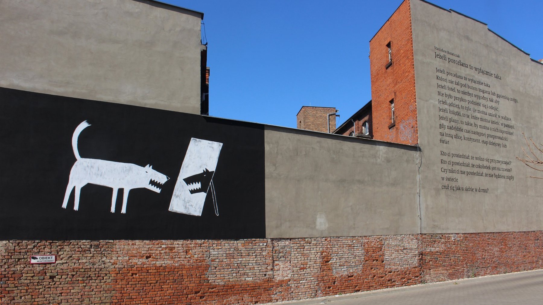 Boczna ściana kamienicy. Czarno-biały mural z psem, który patrzy w lustro. Obok mural z wierszem Stanisława Barańczaka.