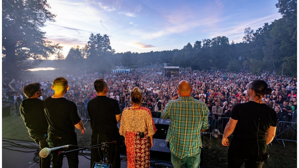 Członkowie zespołu Mikromusic stoją na scenie przed publicznością. Są zwróceni tyłem do obiektywu. W tle widać jezioro.