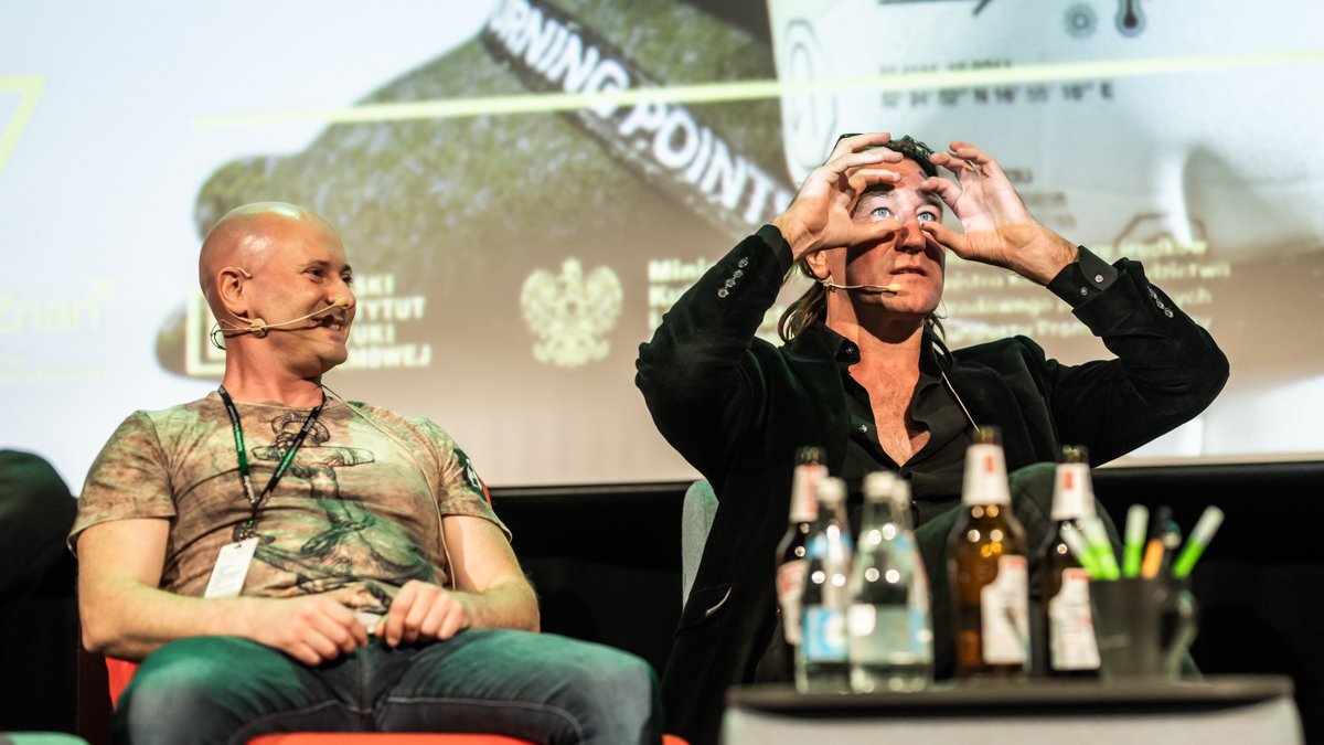 Dwóch mężczyzn siedzi na scenie na fotelach. Mają przyczepione mikrofony i rozmawiają. W tle prezentacja. Przed mężczyznami stoi stolik z napojami.