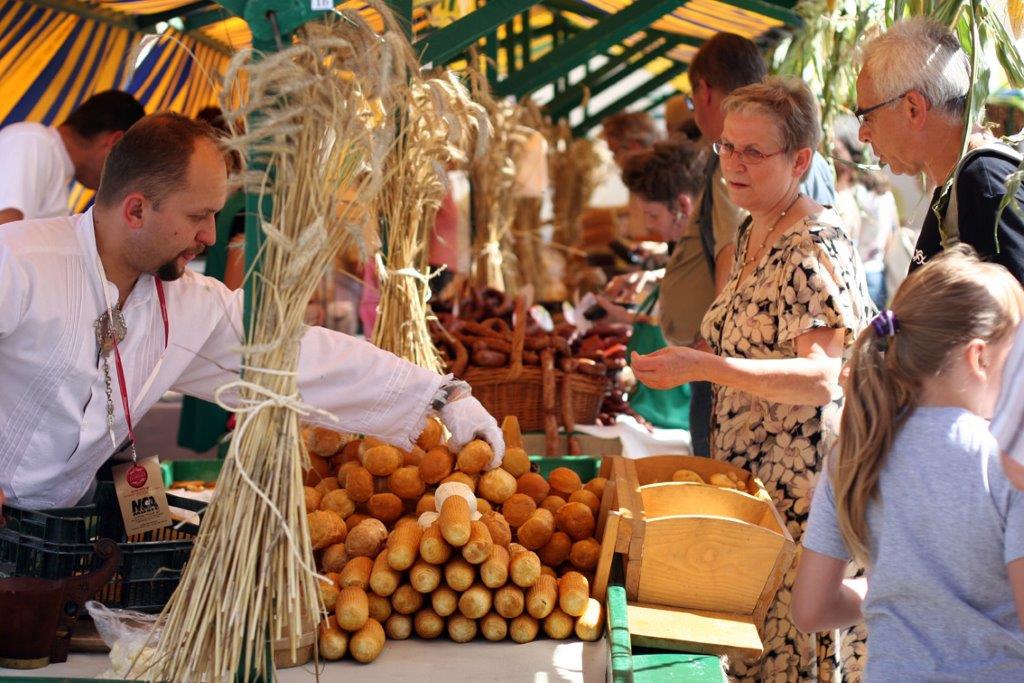 Stragan na Festiwalu Smaków, sprzedawca w białej koszuli oraz klienci, którzy oglądają towary - sery. Stragan udekorowano suszonymi kłosami zboża. - grafika artykułu