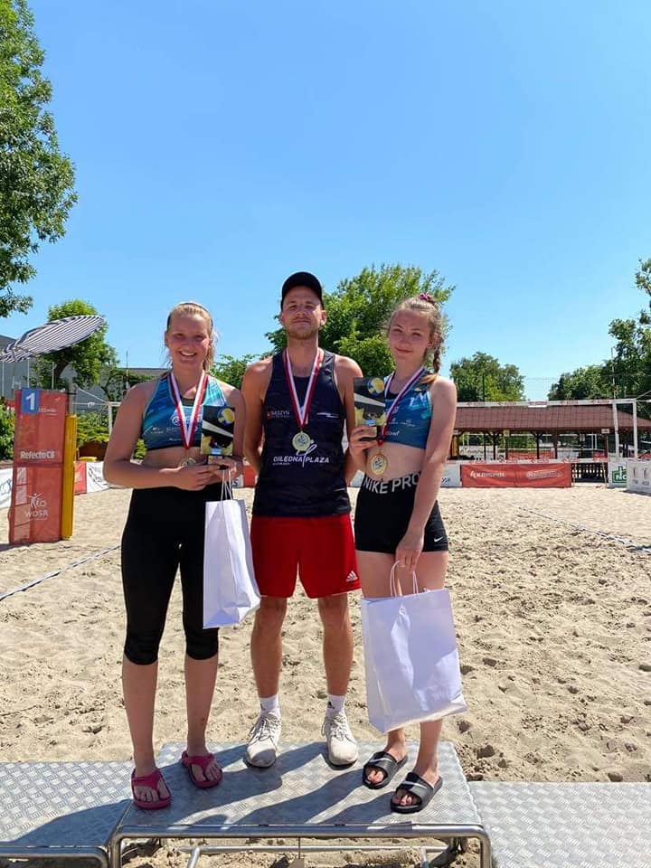 dwie zawodniczki oraz zawodnik stojący pośrodku na tle boiska do siatkówki plażowej - grafika artykułu