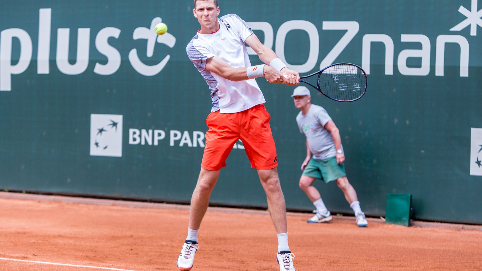 Hubert Hurkacz podczas turnieju Enea Poznań Open, fot. Paweł Rychter - zawodnik w białej koszulce i czerwonych spodenkach szykuje się do odebrania backhandem lecącej piłki. W tle widać sędziego oraz zielony baner z logotypami sponsorów.