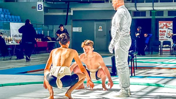 Zawodnicy Sobieskiego na MP seniorów i juniorów w sumo, fot. Jakub Tim - 2 zawodników kuca naprzeciwko siebie, mając tylko przepaskę biodrową i spodenki. Obok stoi sędzia w białym stroju. W tle widać stojących przemieszczających się ludzi, drzwi, puste krzesłeka oraz puchary