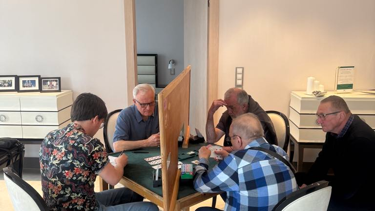 Rozgrywki play off o awans do Brydżowej Ekstraklasy, fot. City Park Bridge Club - 4 mężczyzn siedzi przy stole i gra w karty, jeszcze jeden mężczyzna siedzi i przypatruje się grze