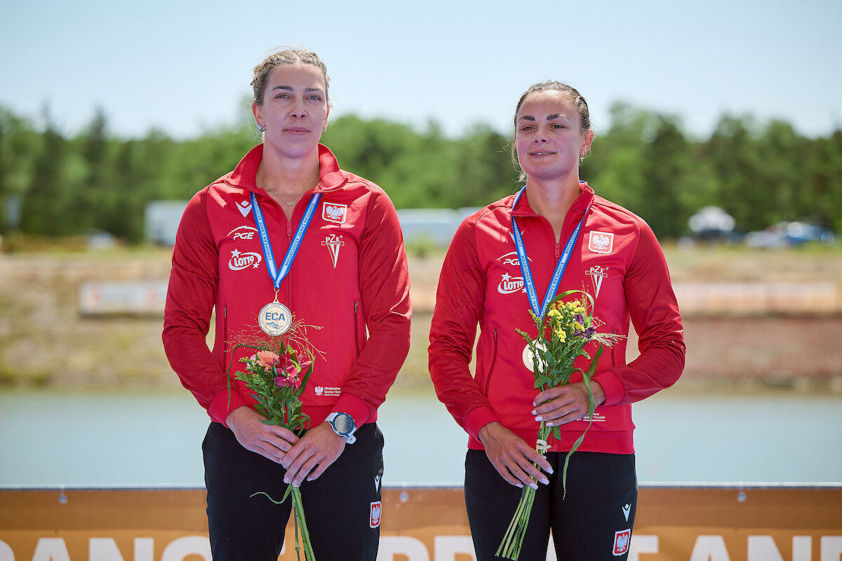 Martyna Klatt i Anna Puławska ze złotymi medalami ME w kajakarstwie, fot. Facebook Polski Związek Kajakowy/Polish Canoe Federation - 2 zawodniczki w czerwonych bluzach i czarnych spodniach stoją z medalami na szyi i kwiatami w rękach - grafika artykułu