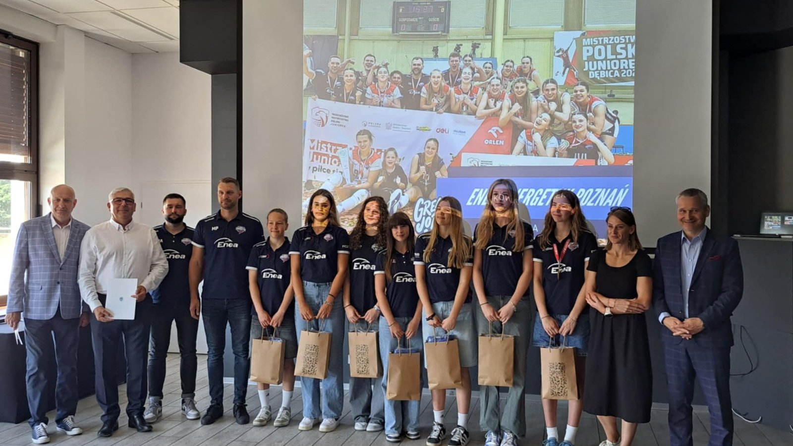 Spotkanie z Młodzieżowymi Mistrzami Polski - z upominkami stoją zawodniczki w ciemnych strojach klubowych. Po bokach stoją przedstawiciele sztabu trenerskiego i osoby wręczające nagrody z ramienia Miasta Poznania