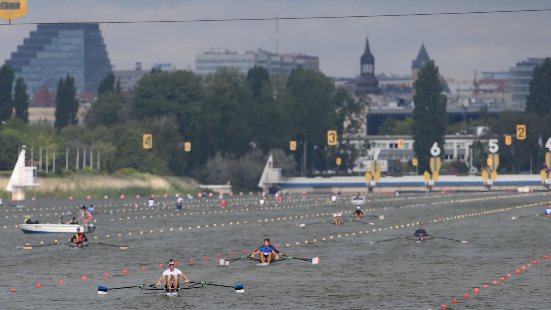 Wioślarze na Torze Regatowym Malta, fot. materiały prasowe OOM - na torze regatowym płynie kilku wioślarzy. Tor jest przygotowany do zawodów, a w tle widać budynki nowoczesne i zabytki miasta oraz ciemne niebo - grafika artykułu