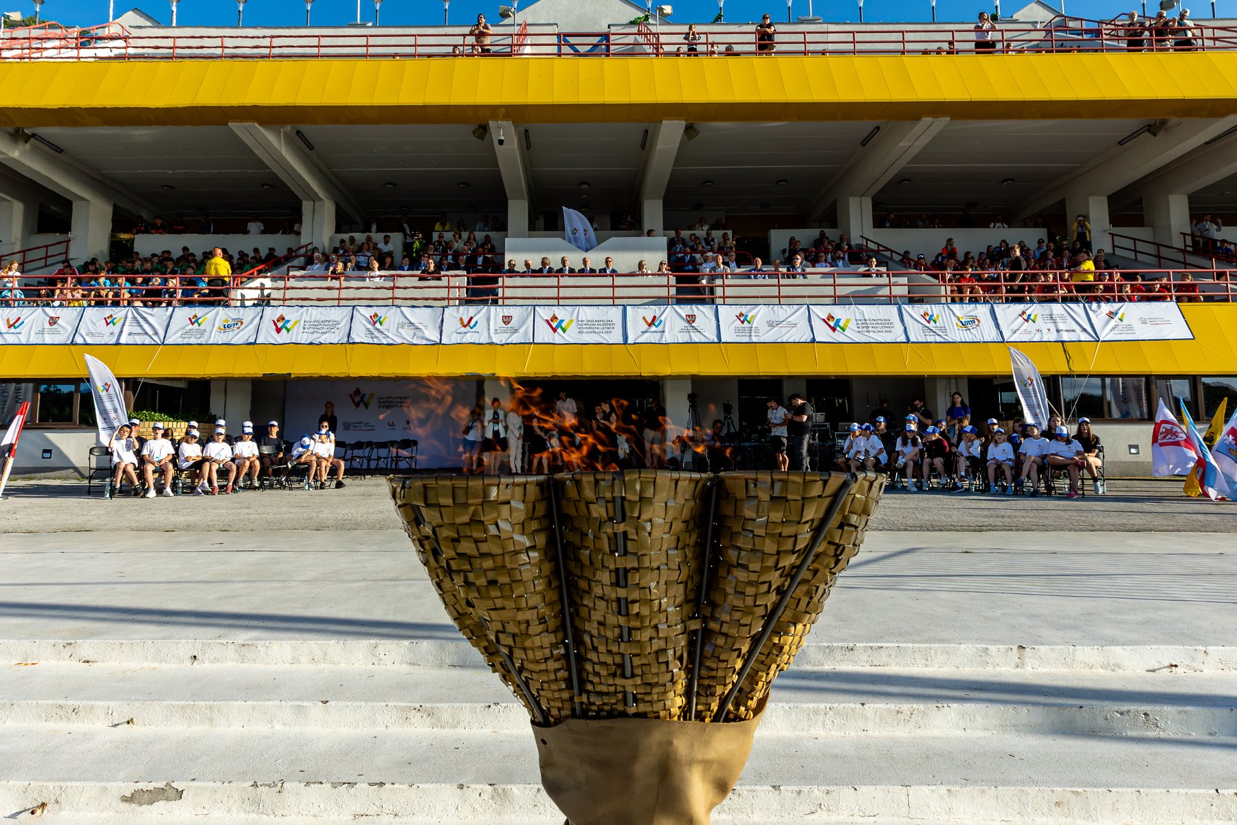 Ogólnopolska Olimpiada Młodzieży 2025, fot. Paweł Rychter - na pierwszym planie płonący znicz, a w tle trybuny i zawodnicy podczas otwarcia zawodów - grafika artykułu