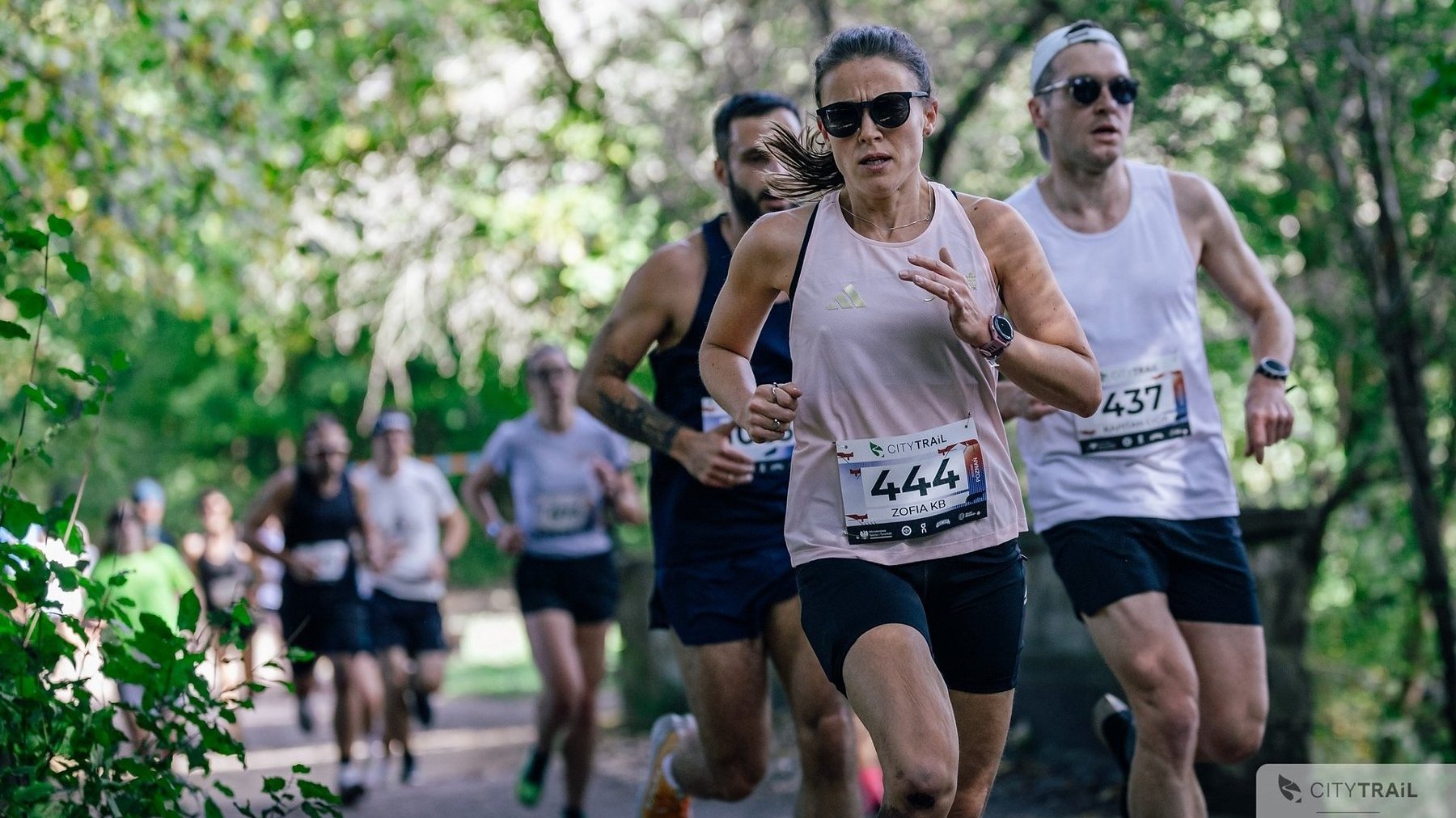 Cykl City Trail w Poznaniu, fot. Andrzej Olszanowski - na pierwszym planie trójka biegaczy w krótkich spodniach i koszulkach (1 kobieta oraz 2 mężczyzn) w tle kilkoro kolejnych biegaczy, a także drzewa z zielonymi liśćmi