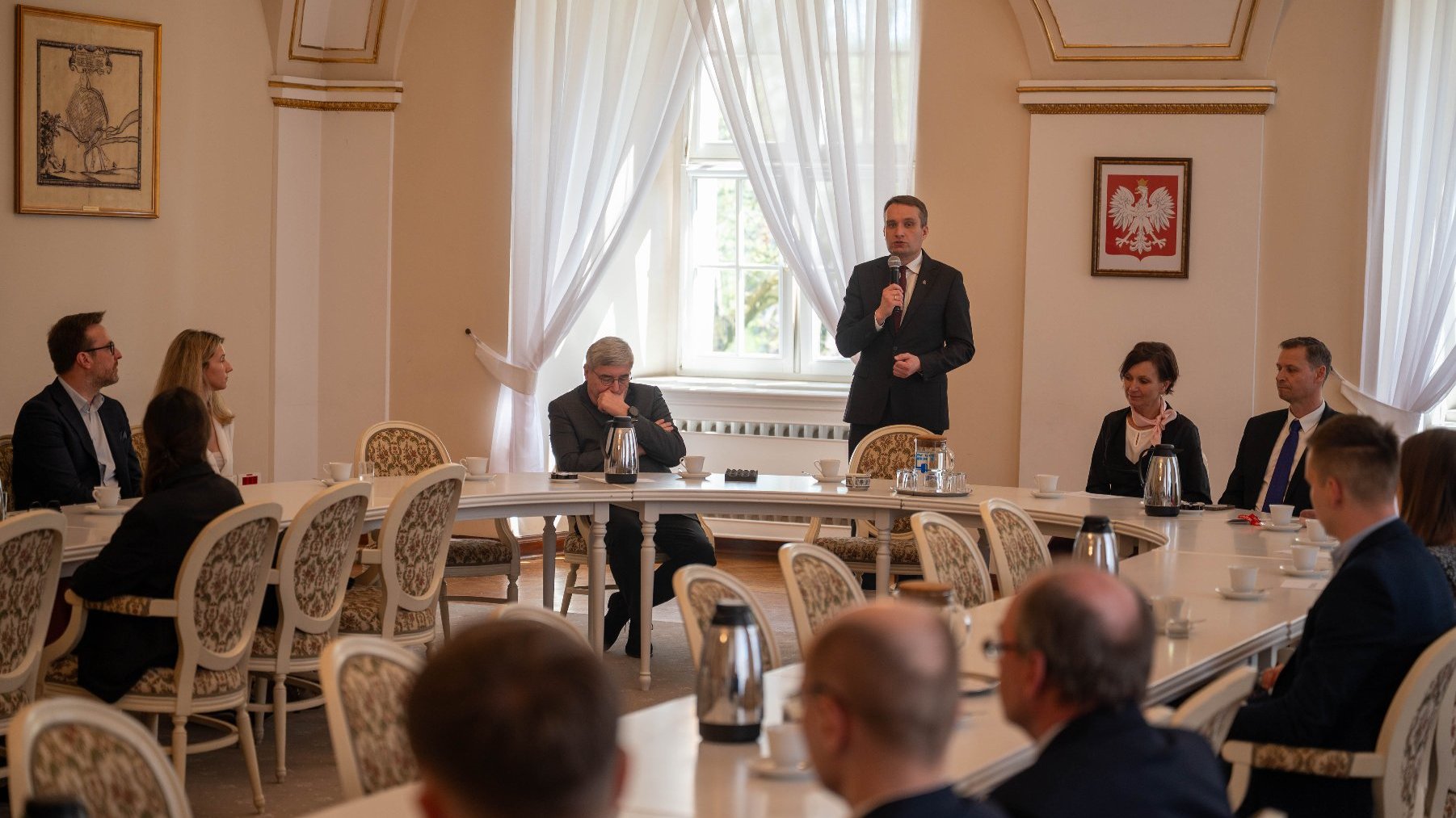 Spotkanie formalne w eleganckim wnętrzu z łukowymi sufitami i dużymi oknami z białymi zasłonami. Przy długim stole siedzi kilka osób, jedna osoba stoi i mówi do mikrofonu. Na