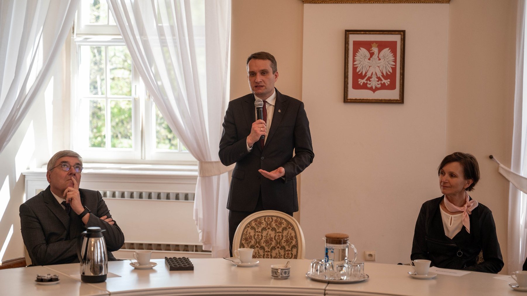 Trzy osoby przy zakrzywionym stole w formalnym otoczeniu.