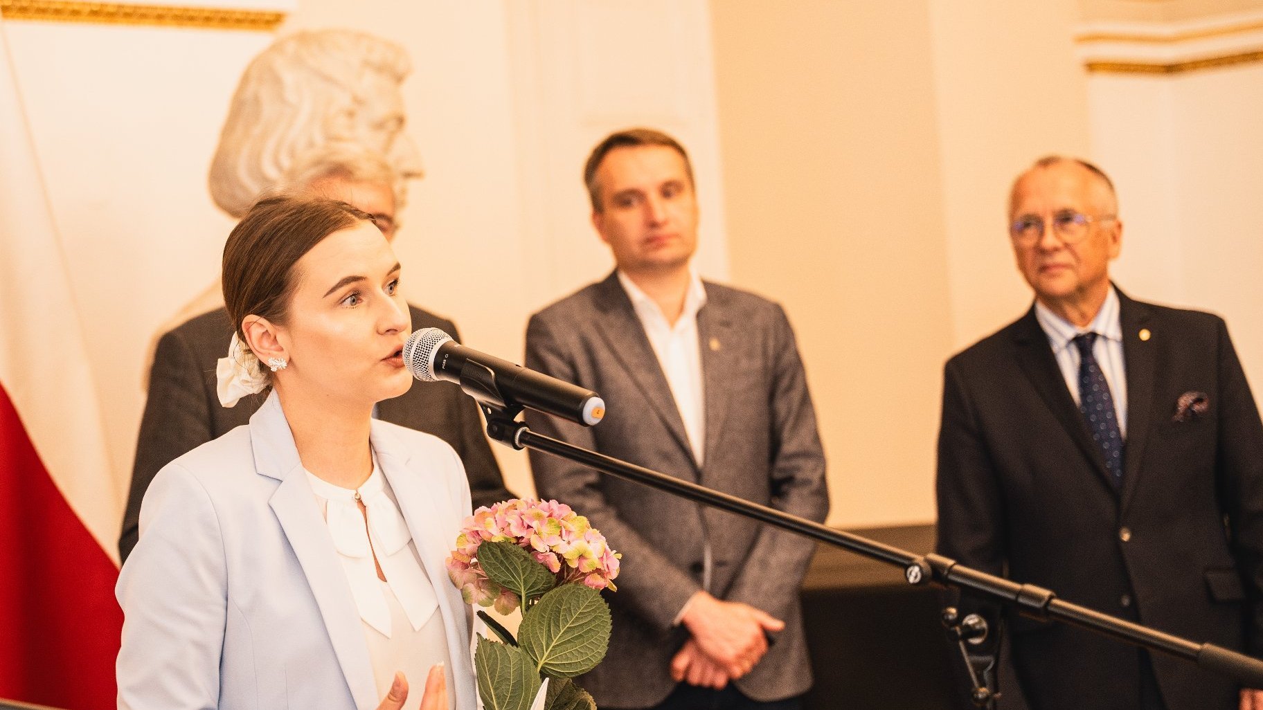 w tle trzech mężczyzn i przed nimi kobieta która mówi przez mikrofon