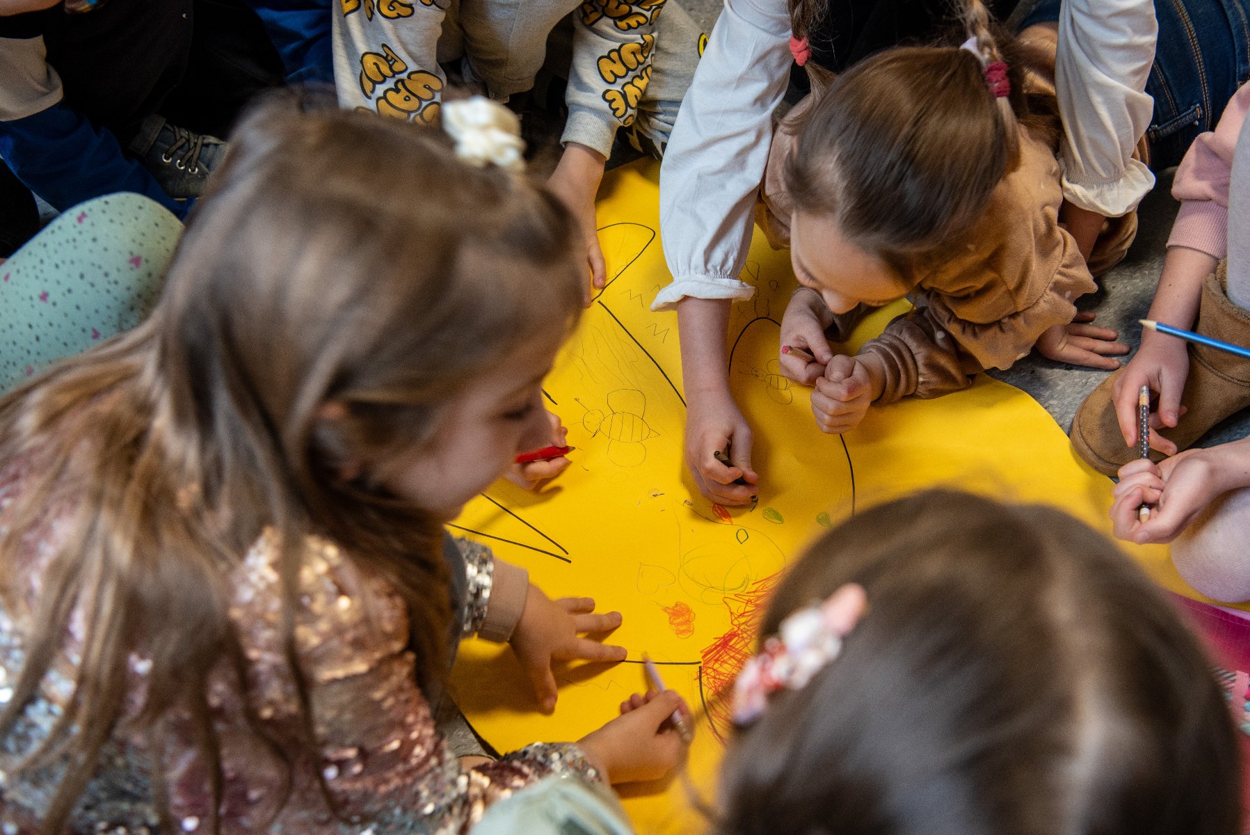 Grupa dzieci rysuje na dużym żółtym arkuszu papieru. Na środku kartki znajduje się szkic przedstawiający kształt przypominający zwierzę lub postać. Wokół szkicu dzieci tworzą mniejsze rysunki i bazgroły. - grafika artykułu