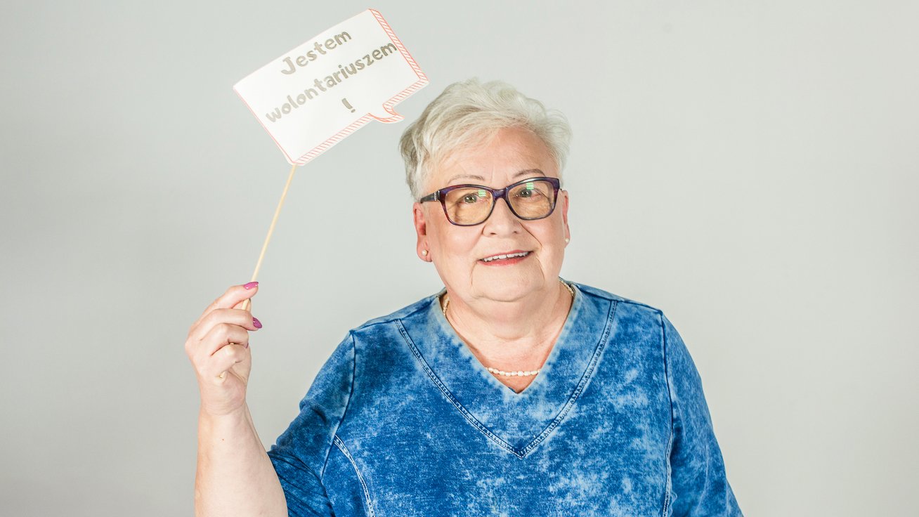 Na zdjęciu starsza kobieta trzymająca tabliczkę z napisem: jestem wolontariuszem