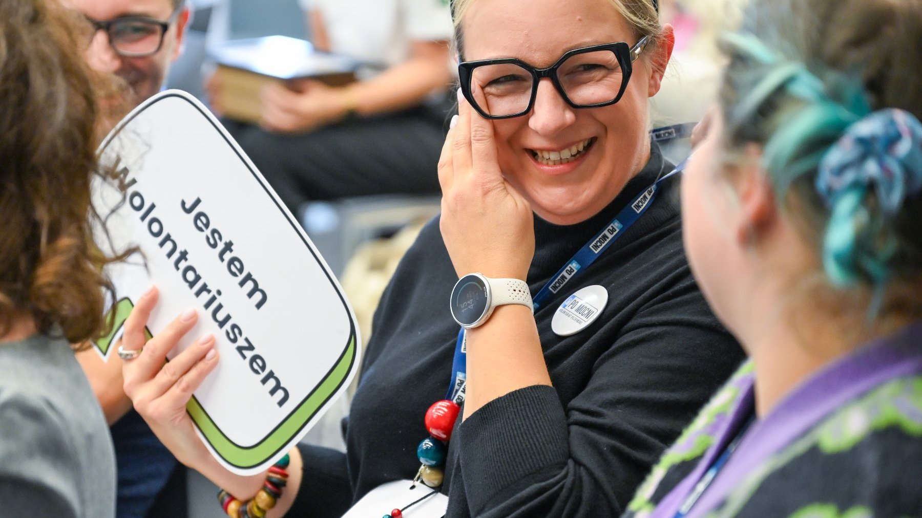 Na zdjęciu dwie uśmiechnięte kobiety, jedna trzyma tabliczkę z napisem: jestem wolontariuszem