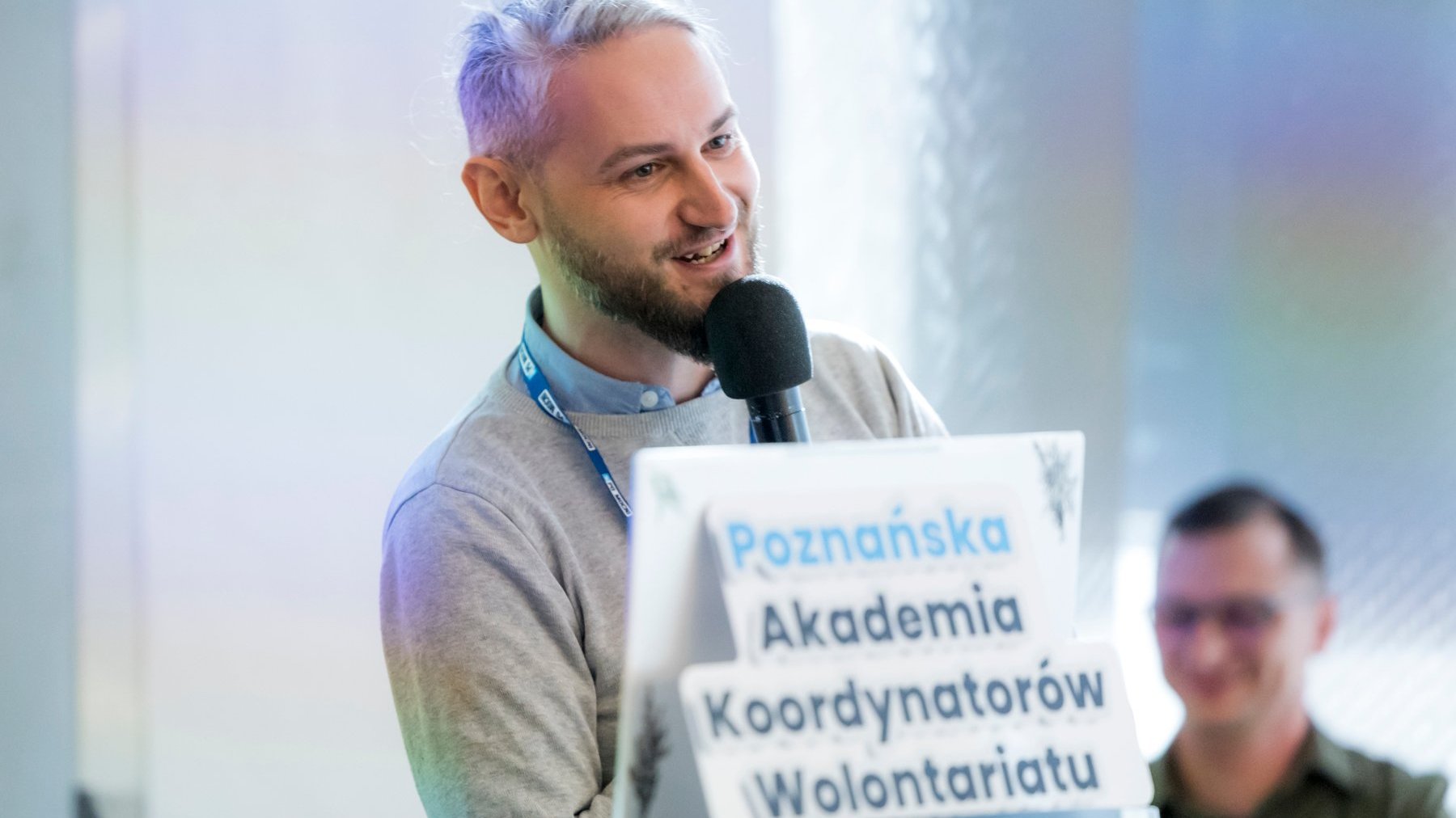 Na zdjęciu mężczyzna z mikrofonem w ręku, pod nim napis: Poznańska Akademia Koordynatorów Wolontariatu