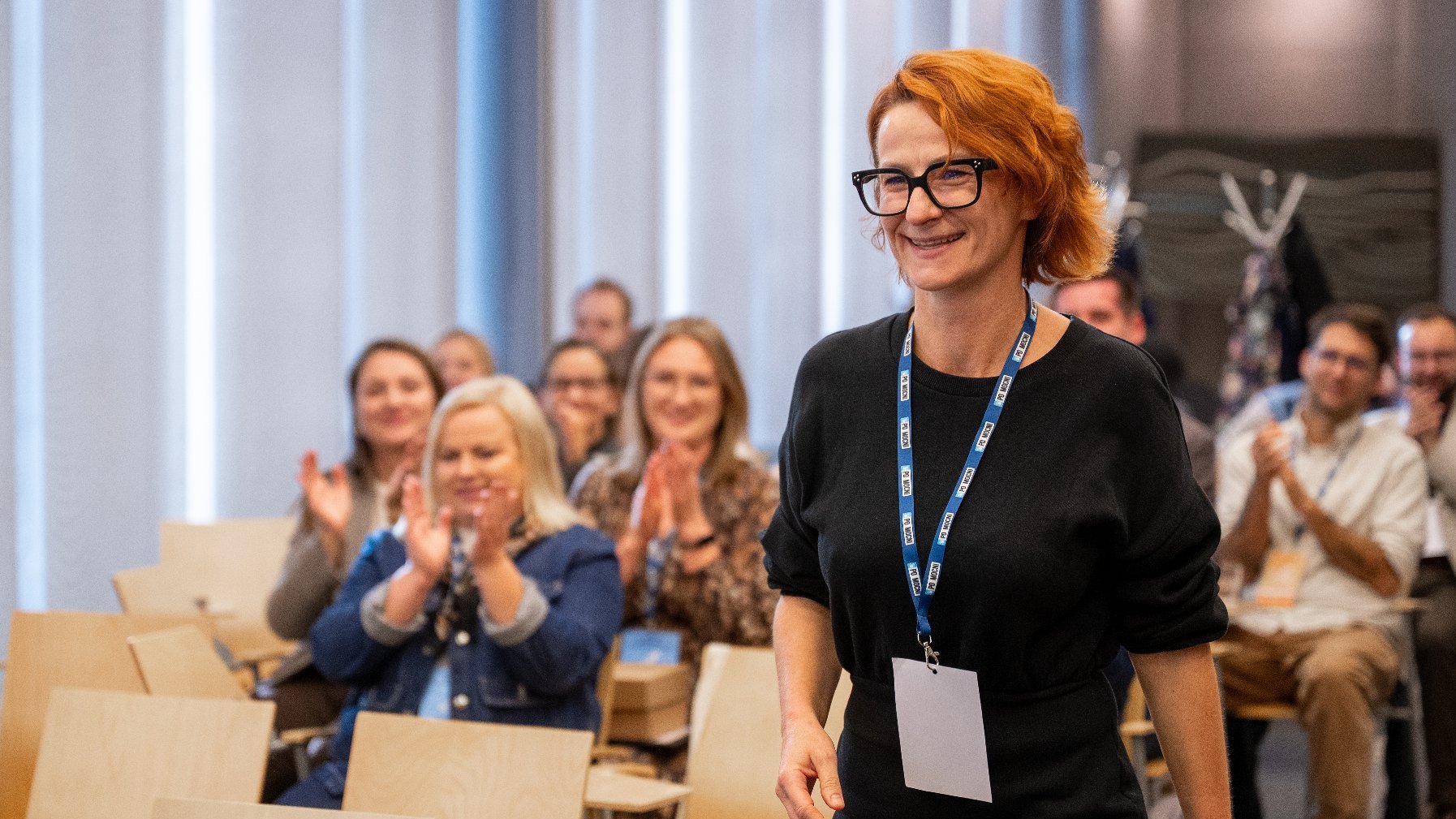 Na zdjęciu uśmiechnięta kobieta, w tle klaszczące postaci